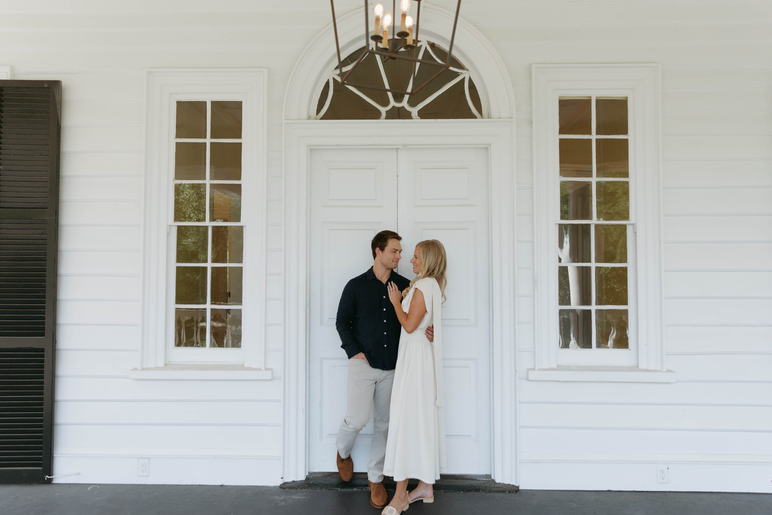 A couple stands close together in front of a white door with large windows, sharing a moment of affection, with a chandelier hanging above them.