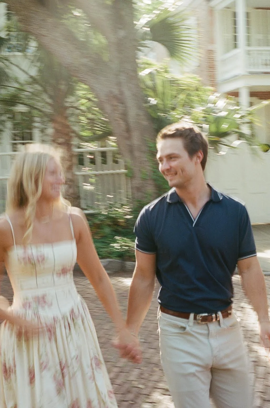 A young couple holding hands and smiling at each other outdoors in a garden or park area with green foliage and a large tree, bathed in warm sunlight.