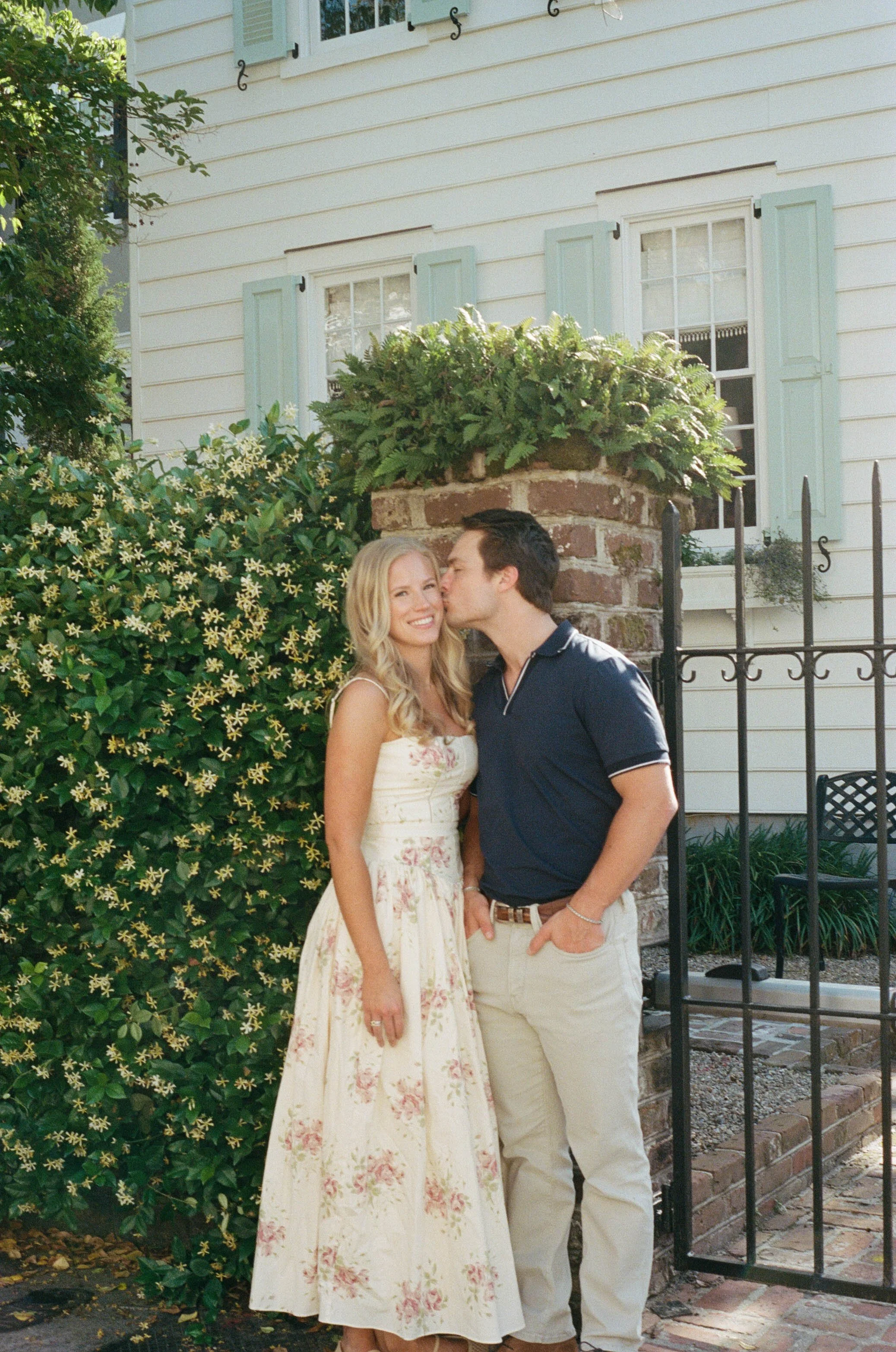 A young woman in a floral dress and a young man in a polo shirt and khakis standing outside in front of a brick pillar with greenery and flowering bushes, with a white house featuring light blue shutters behind them. The man is kissing the woman's ch