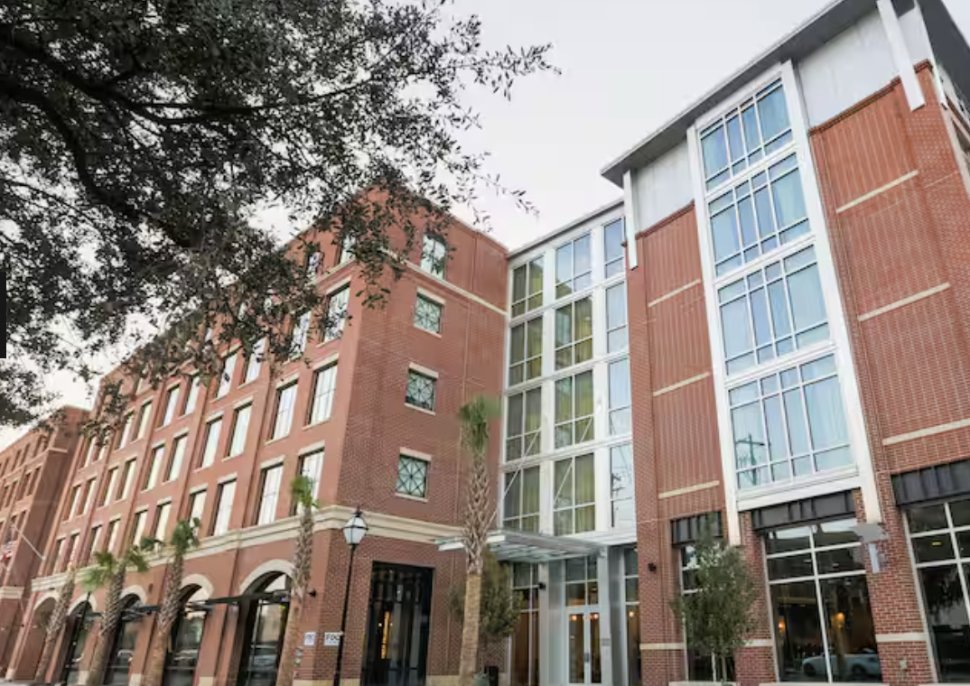 A multi-story building with a brick facade and large glass windows, surrounded by trees with some greenery on the sidewalk.