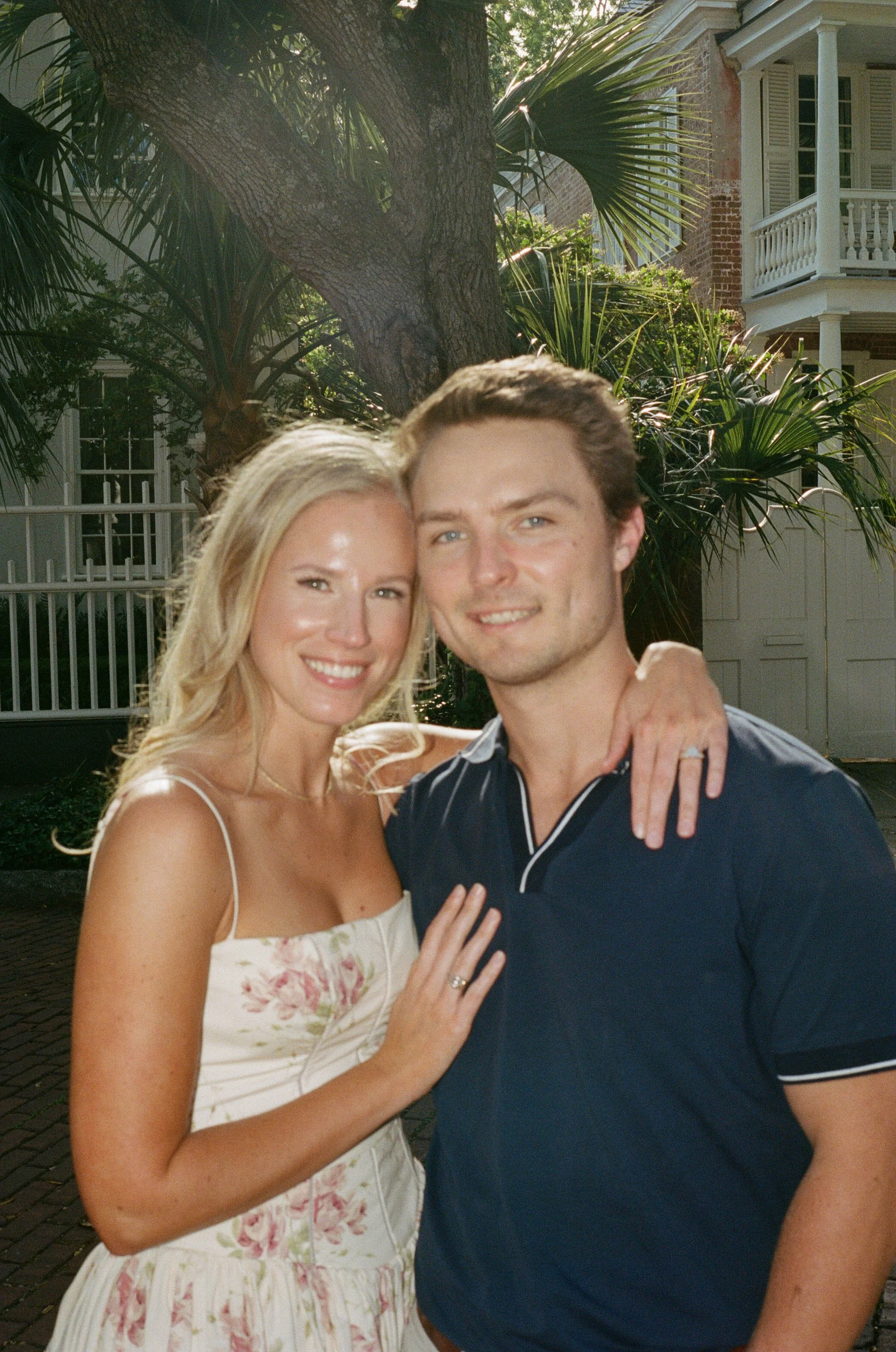 A smiling woman with long blonde hair in a white floral dress and a smiling man with short brown hair wearing a dark shirt are embracing each other outdoors with a large tree, green plants, and a house with a brick facade and white porch in the backg