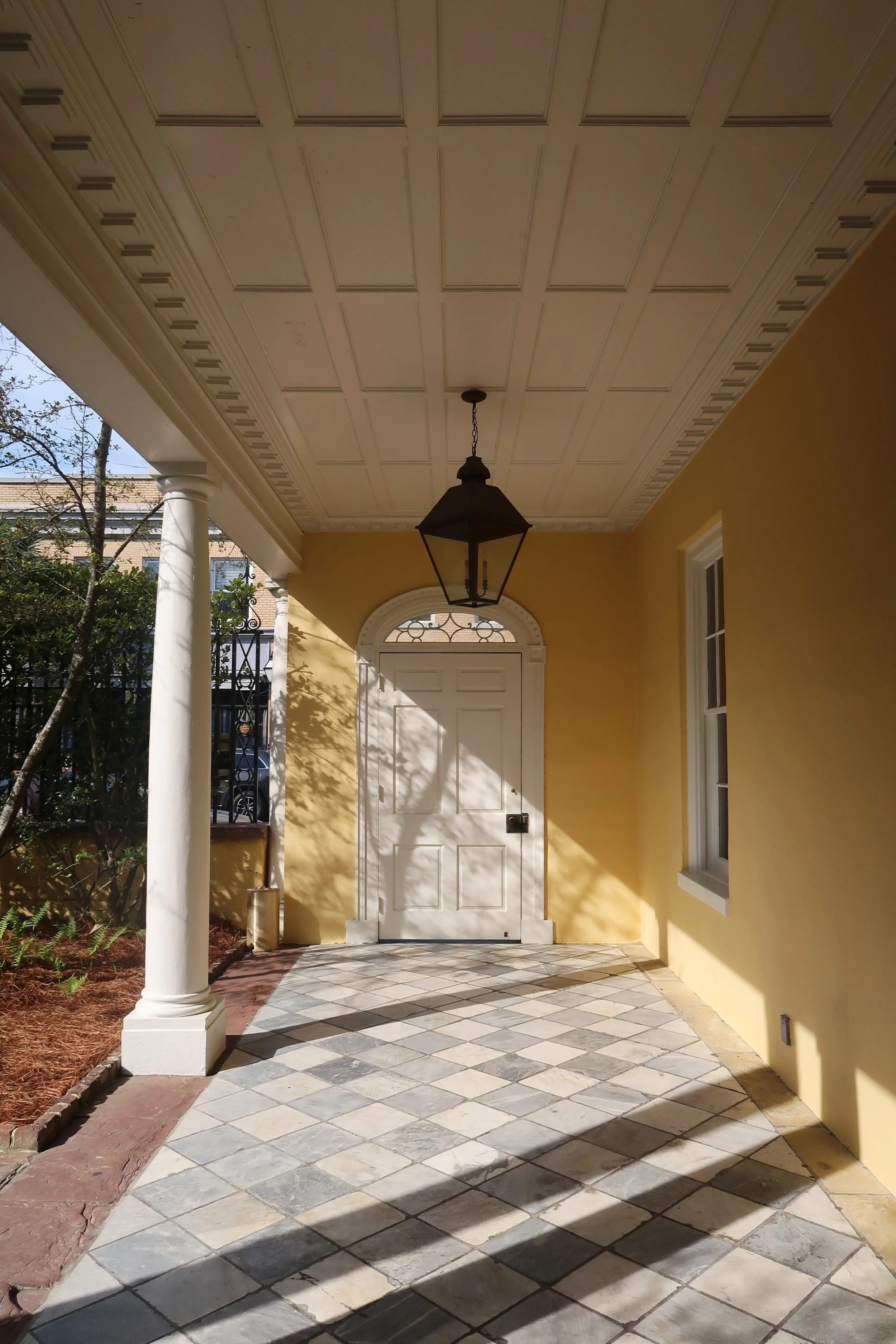 Front porch of a house with yellow walls, white door, hanging lantern, tiled floor, white columns, and landscaping with trees and plants.