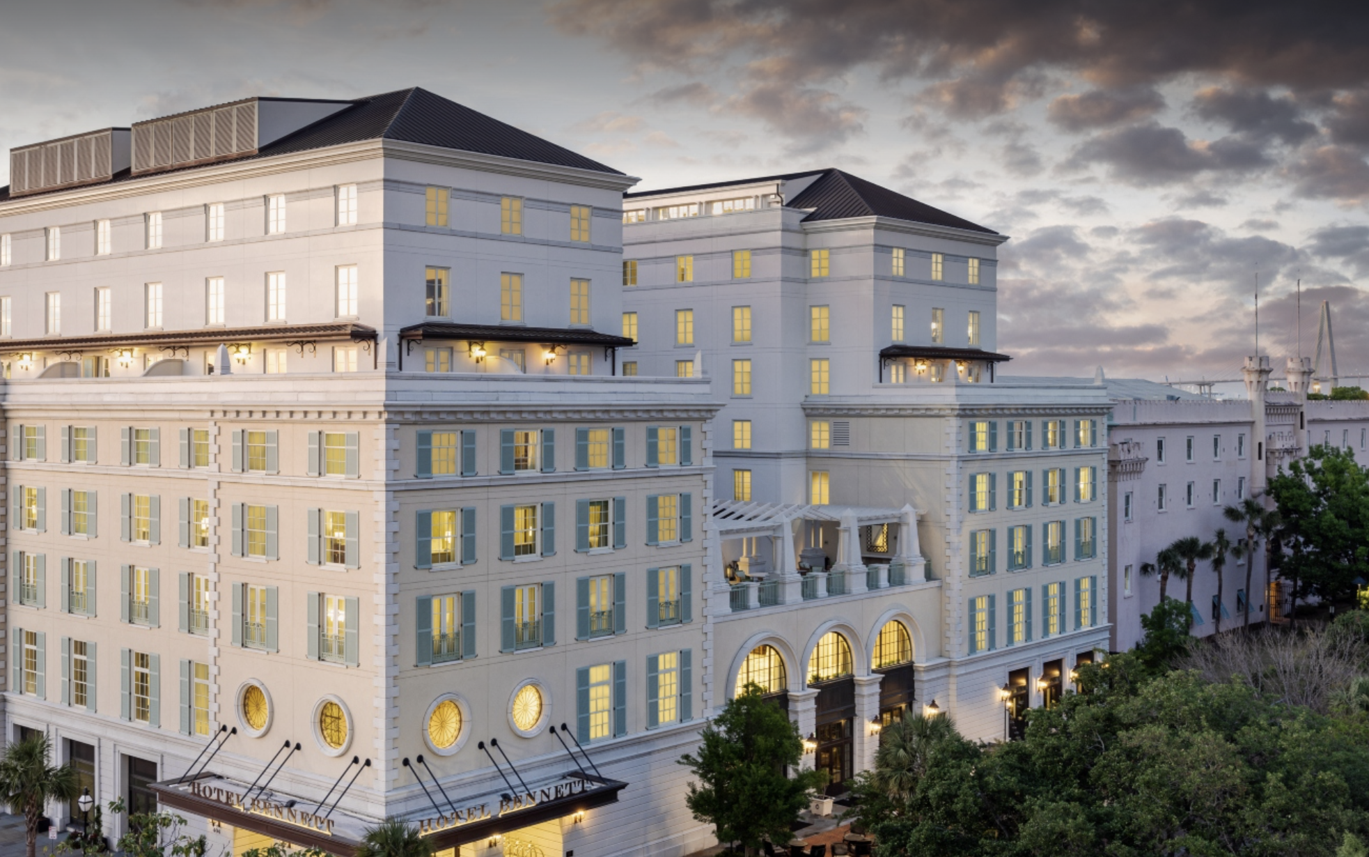 A large white hotel building with numerous lit windows, including a prominent entrance and multiple balconies, set against a partly cloudy sky during dusk.