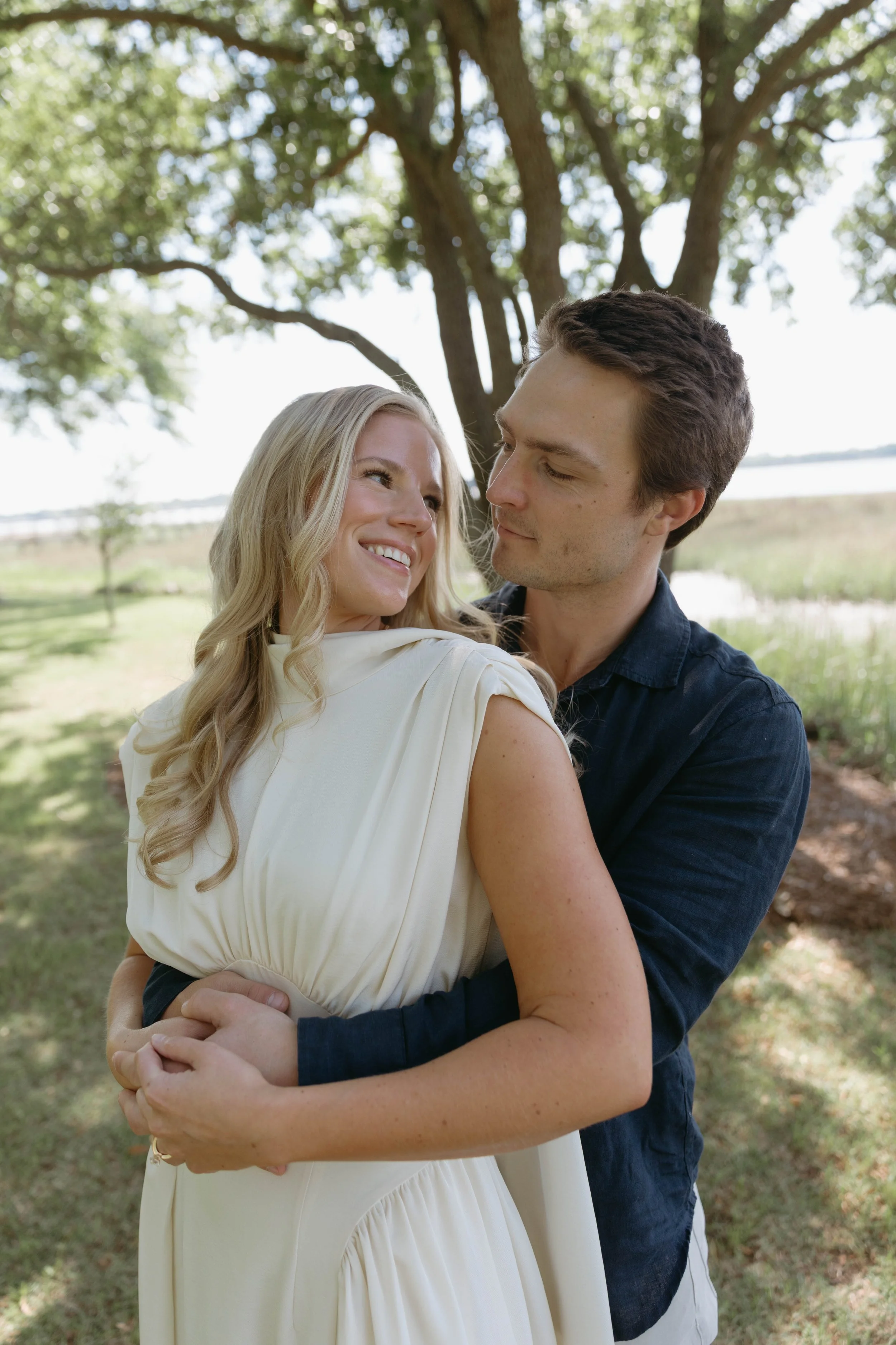 A couple embraces outdoors under a tree, smiling and gazing at each other, with water and greenery in the background.