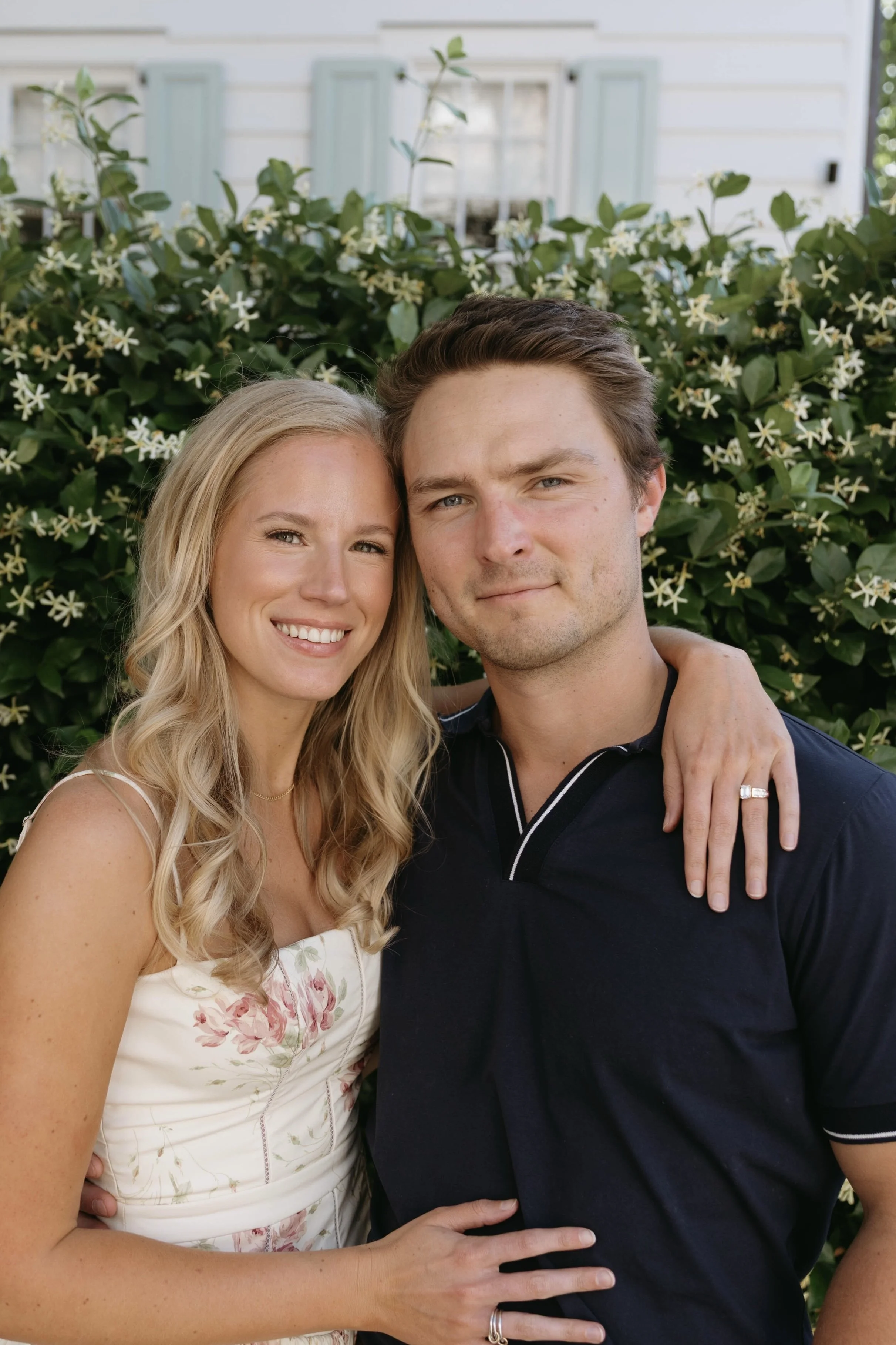 A woman with long blonde hair smiling and a man with short brown hair smiling, standing close together outside in front of a bush with white flowers and a white house with light blue shutters in the background.