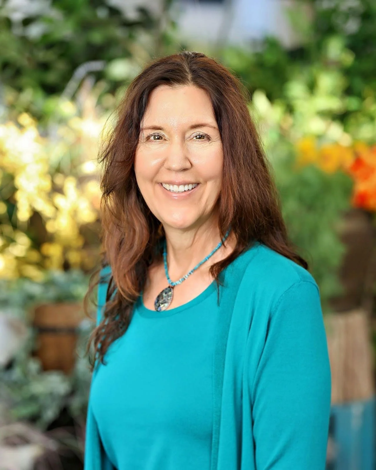 Smiling woman with long brown hair wearing a turquoise top and necklace, standing outdoors with blurred greenery and flowers in the background.