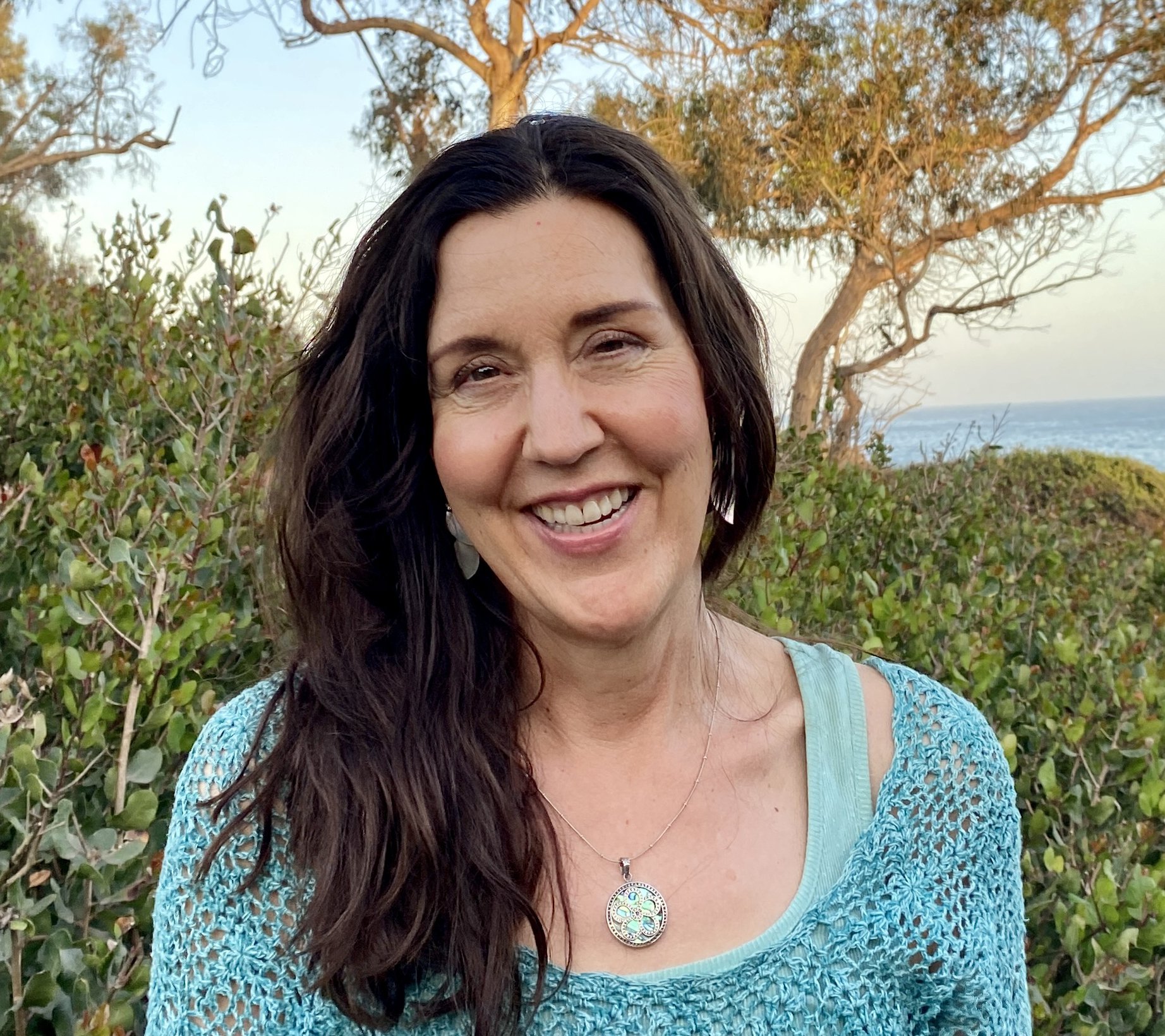 Close-up of a smiling woman with dark hair wearing a turquoise crocheted top and a pendant necklace, standing outdoors with green shrubbery, trees, and the ocean in the background during sunset.