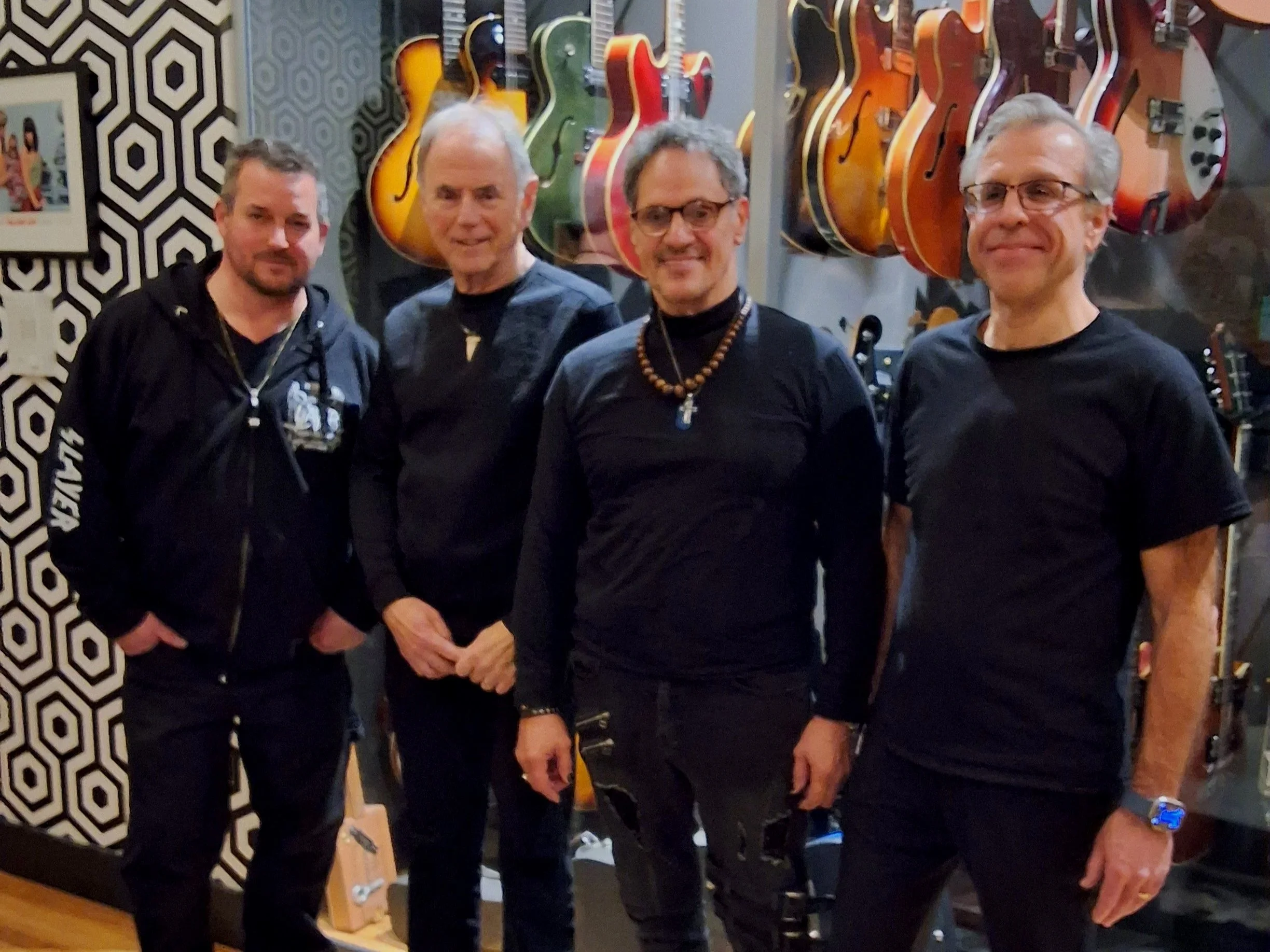 Four men standing in a music store with guitars hanging on the wall behind them.