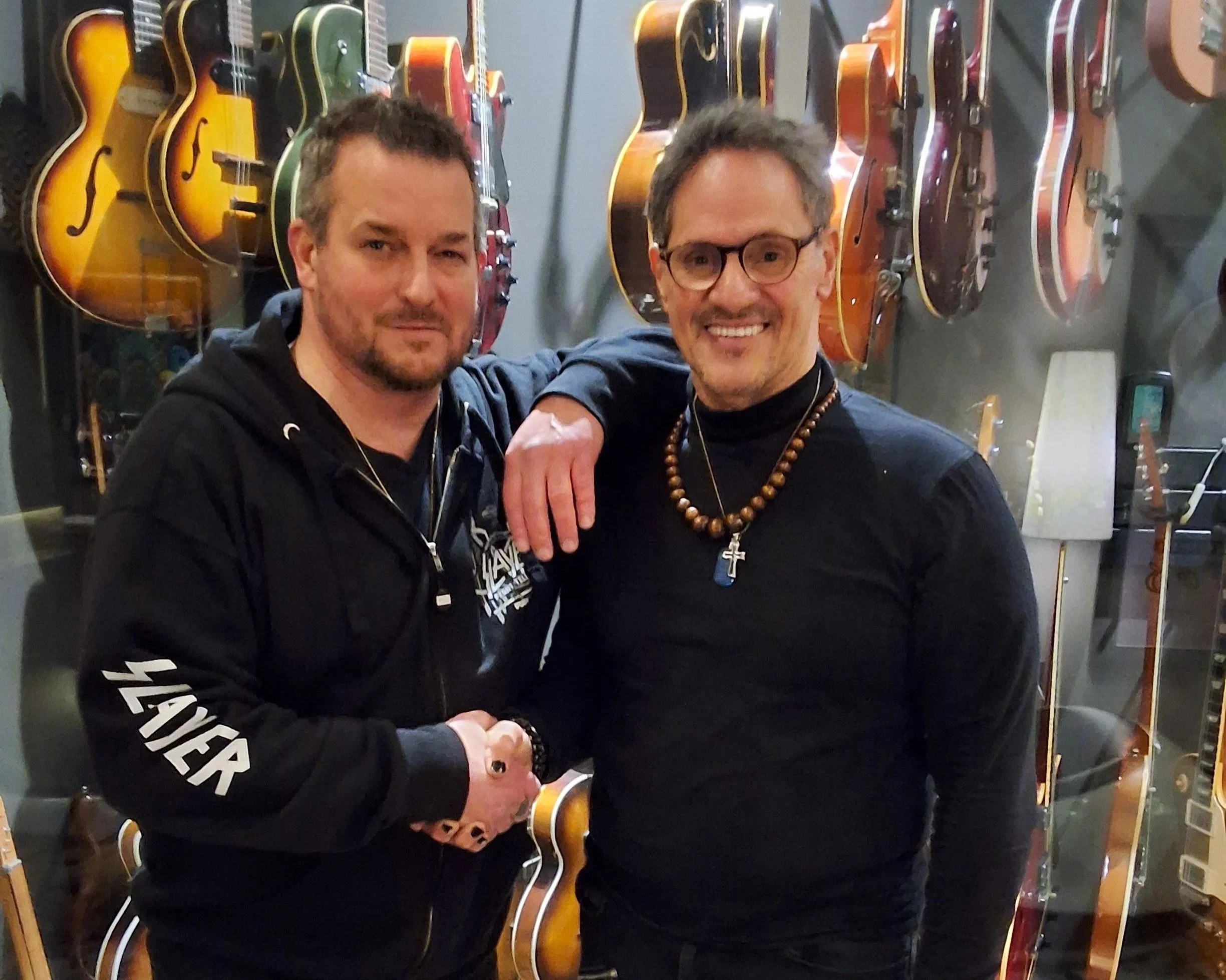 Two men standing in front of a display of various guitars hanging on the wall, shaking hands and smiling.