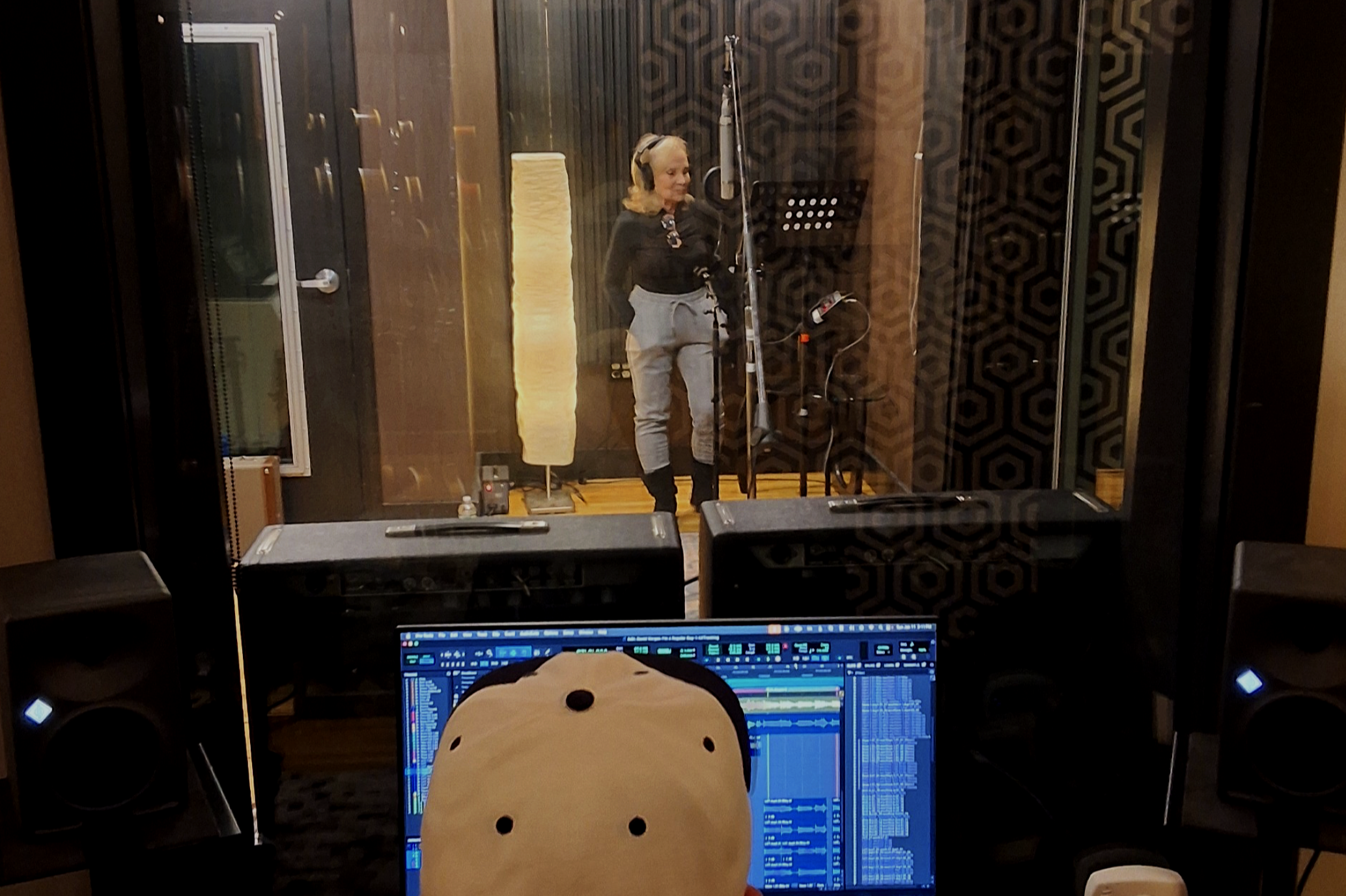 A woman singing into a microphone in a recording studio, seen through a glass window from the control room where a person is working on a computer with music production software.