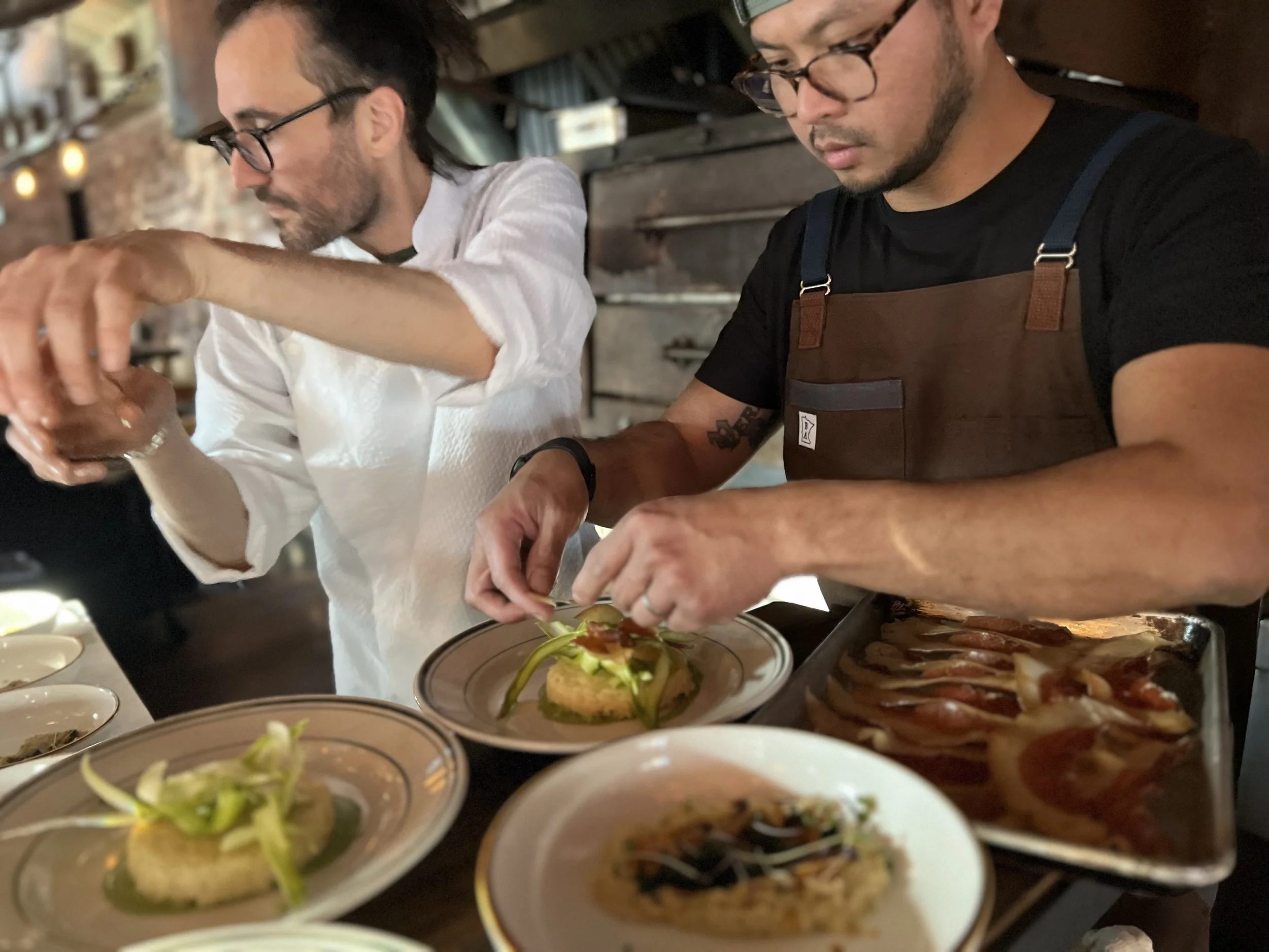 Two male chefs are preparing dishes in a restaurant kitchen. They are plating food and garnishing plates with various ingredients, with one wearing a white chef's coat and the other wearing a brown apron.