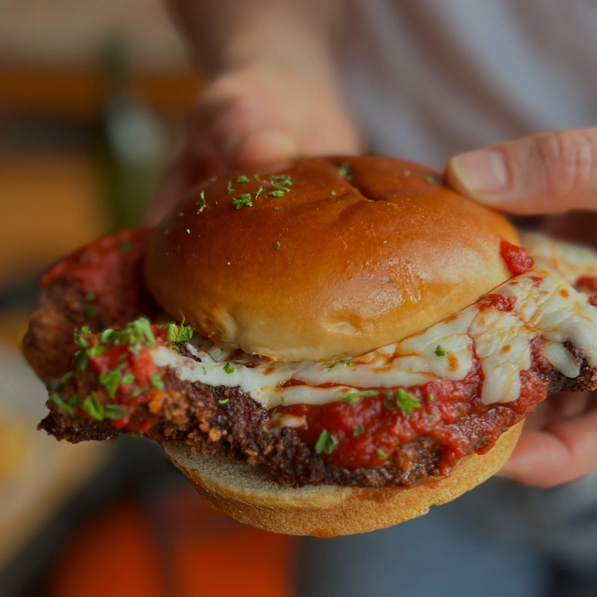 Close-up of a cheeseburger with a beef patty, melted cheese, tomato sauce, and a shiny bun, garnished with chopped herbs, held in a person's hand.