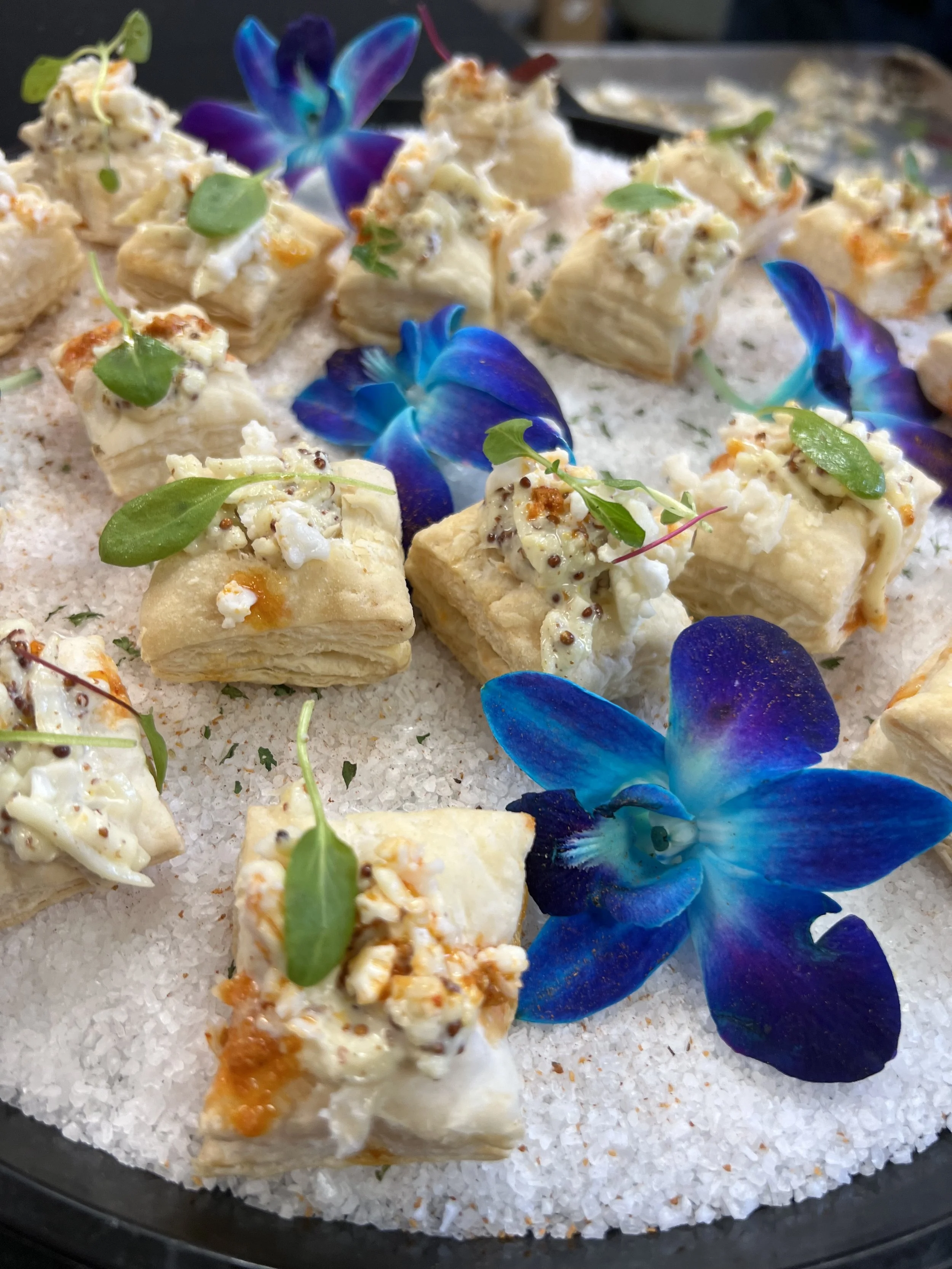 Assorted bites of flaky pastry topped with creamy filling, garnished with microgreens, surrounded by vibrant blue orchid flowers on a bed of coarse white salt.