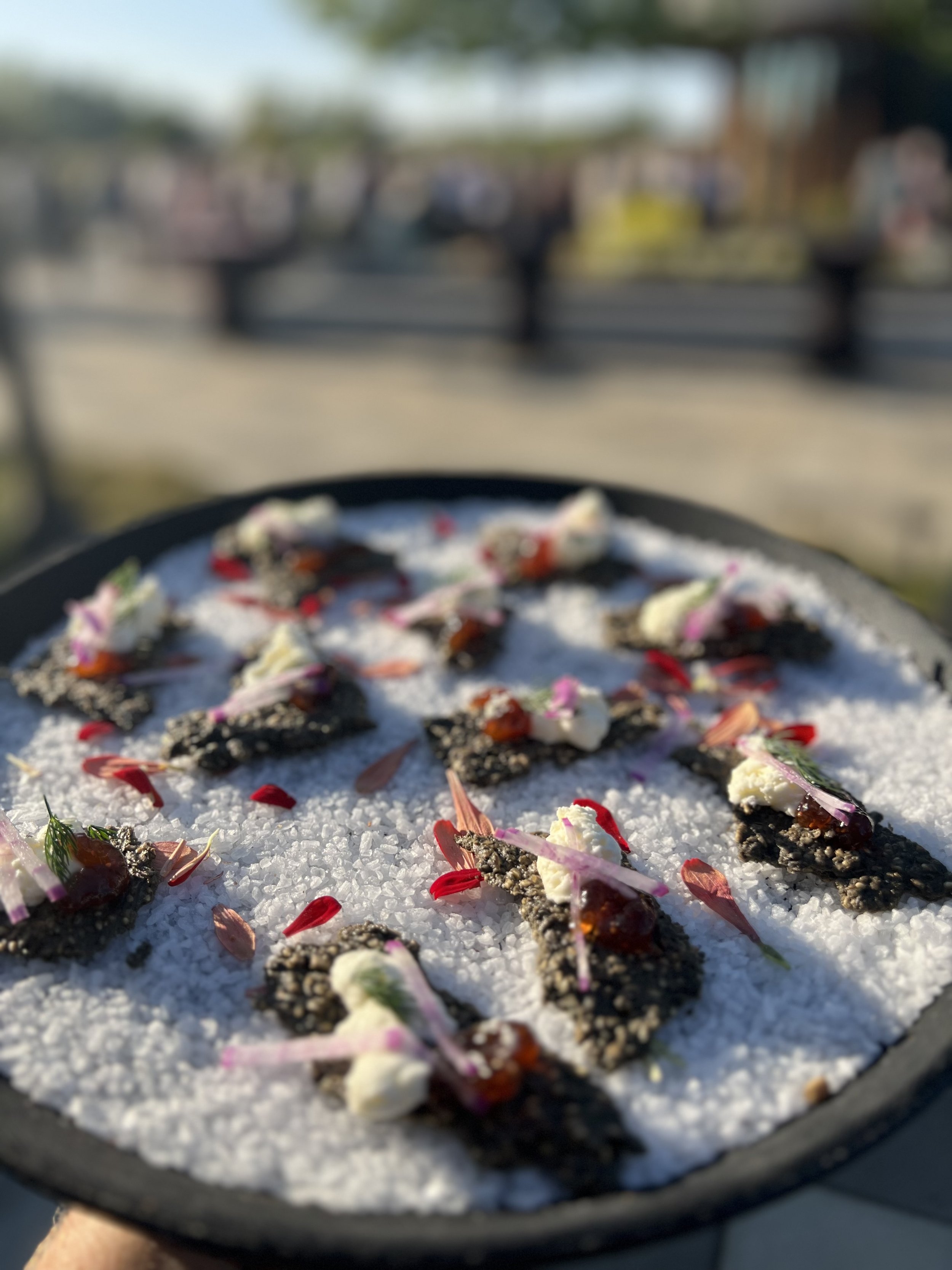 Close-up of a dish with small portions of food garnished with flowers, on a black plate with sea salt, outdoors with a blurred background.