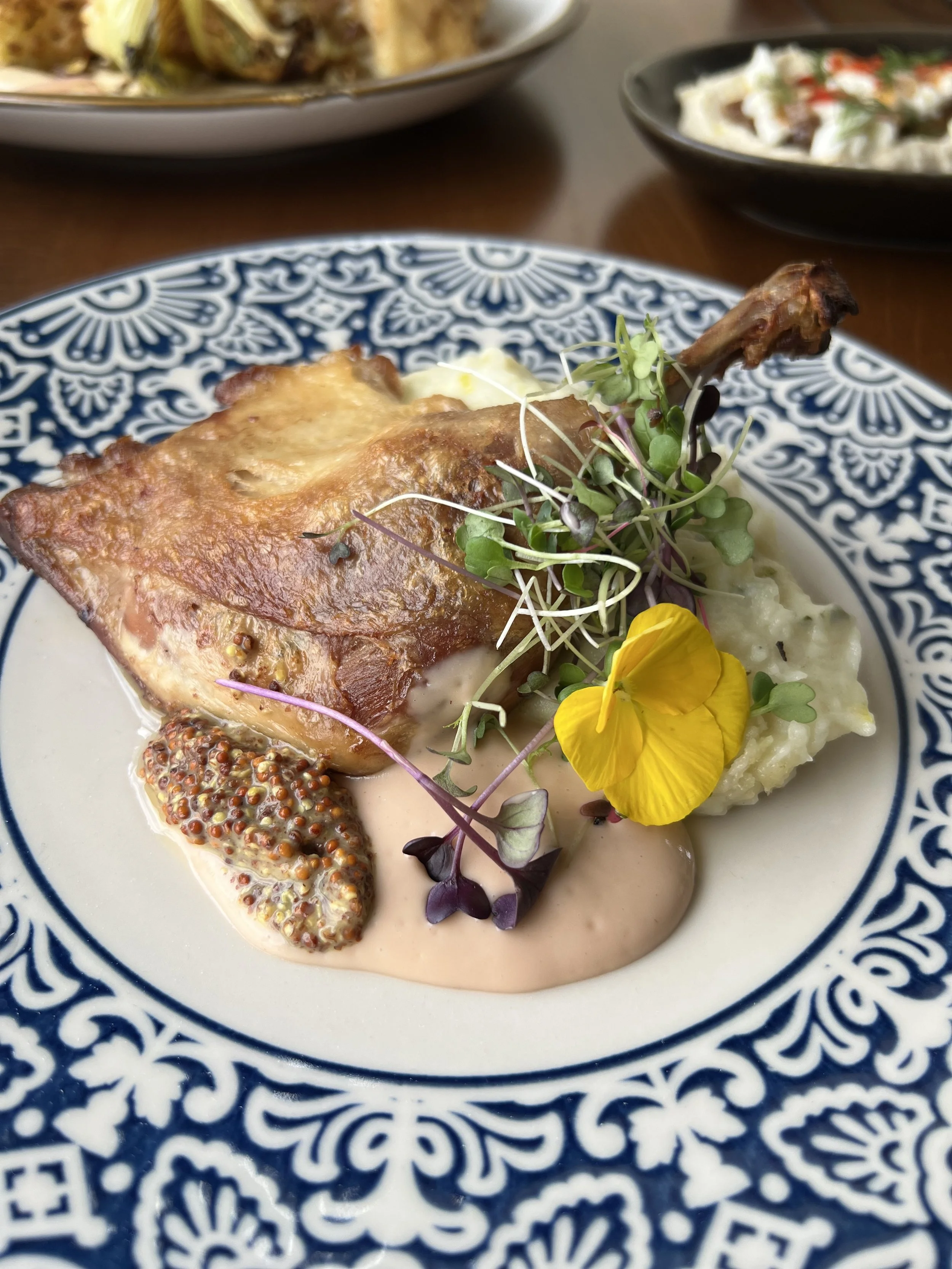 A plate with a cooked chicken leg, potato mash, mustard, microgreens, and a yellow edible flower, garnished with a creamy sauce.