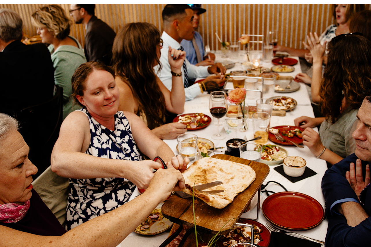 People sitting at a dinner table with plates of food, glasses of wine, and a large piece of bread.