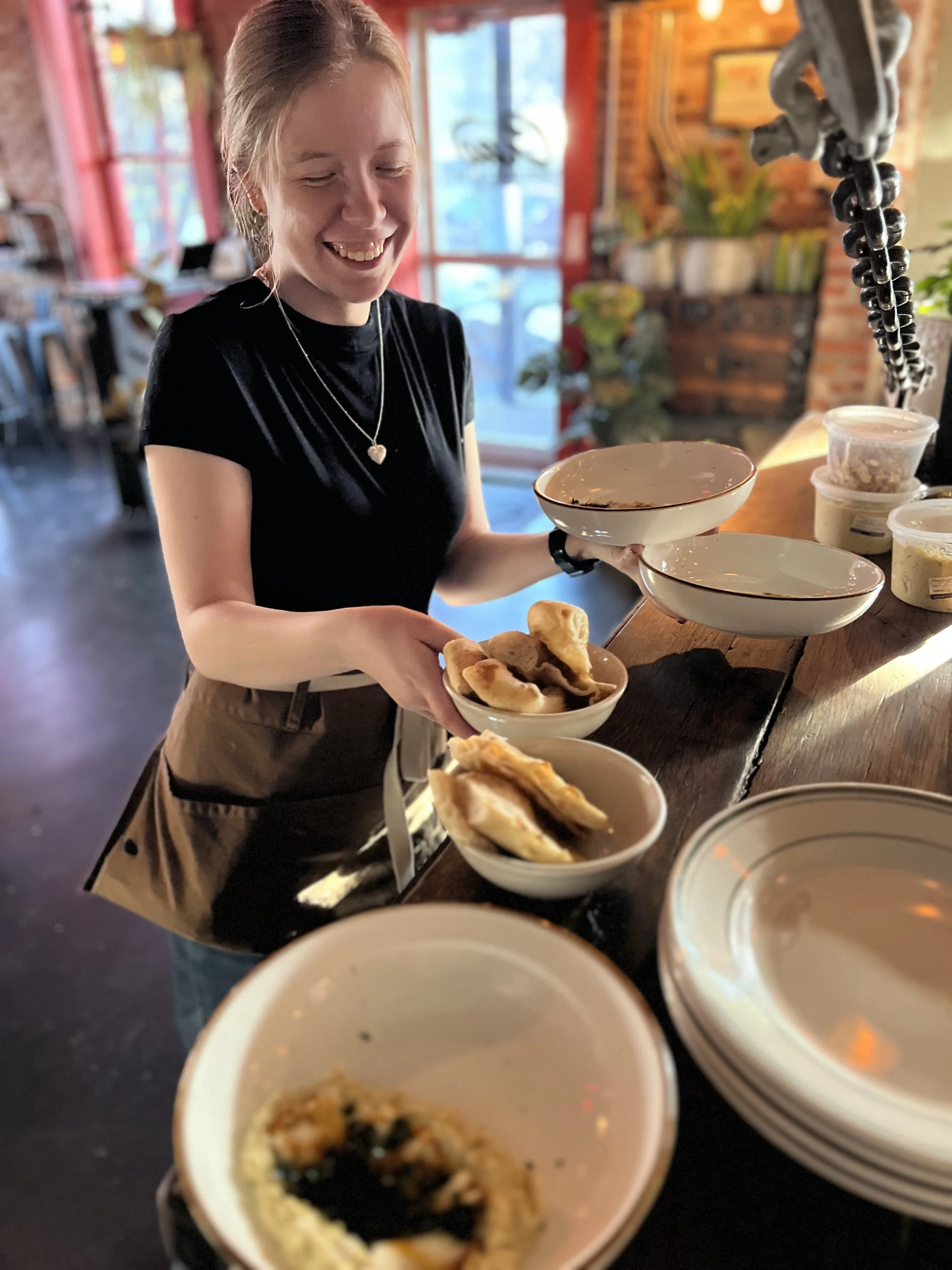 A woman in a black T-shirt and brown apron is smiling while stacking bowls of dumplings in a restaurant with a rustic interior, red window frames, and plants on the counter.