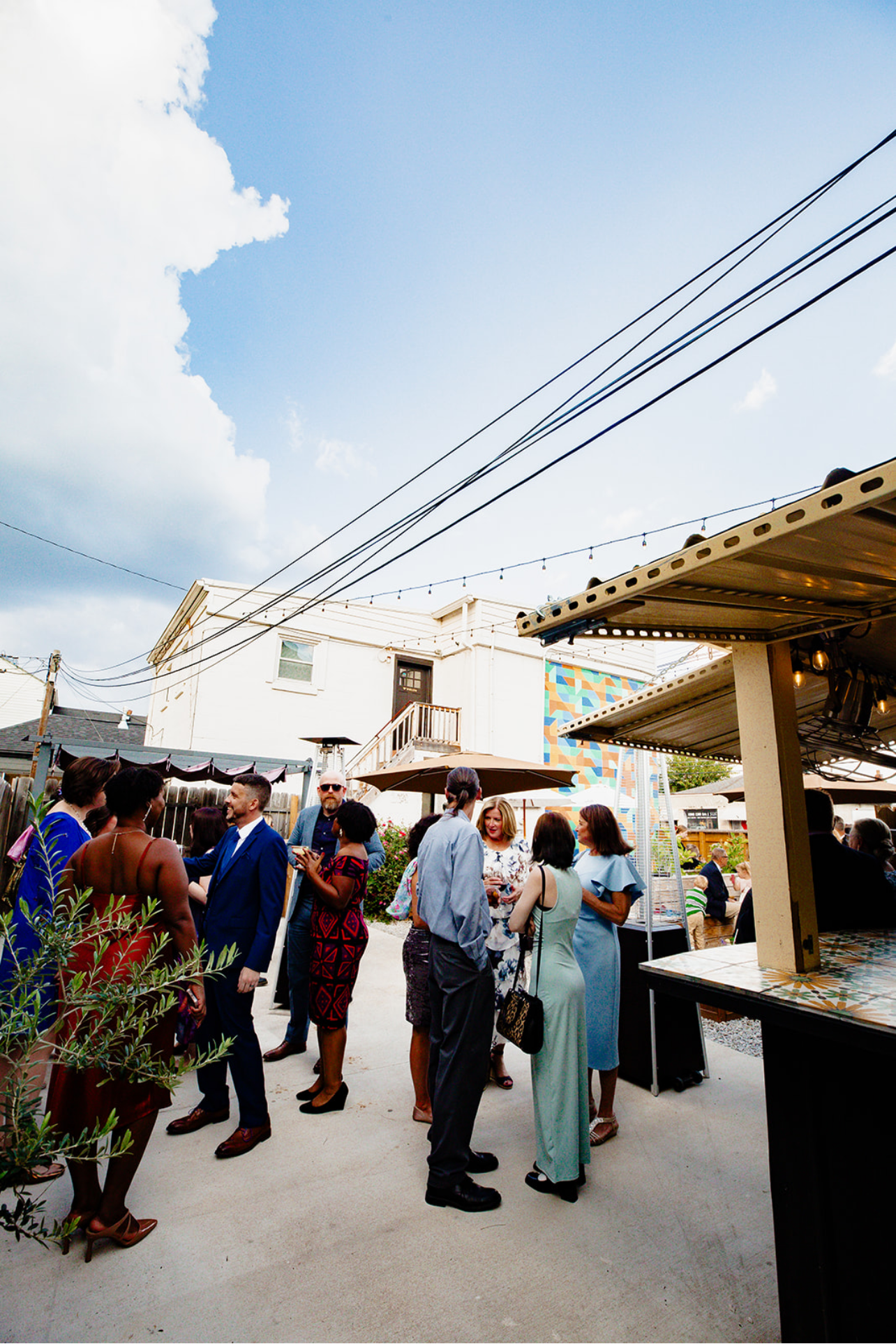 Group of people socializing at an outdoor gathering on a sunny day, with a building, colorful mural, and sky with clouds in the background.