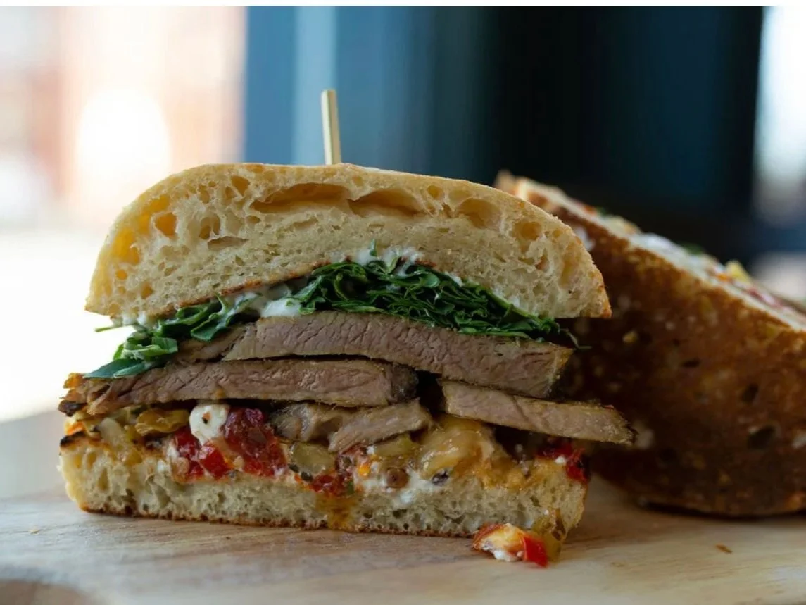Close-up of a sliced sandwich with beef, greens, cheese, and vegetables on a crusty bread roll, on a wooden surface.