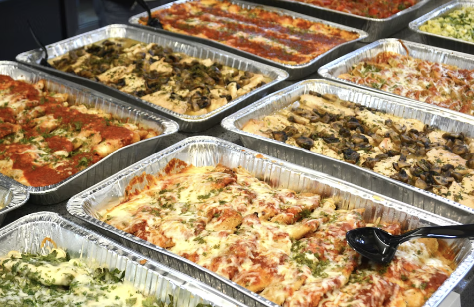Multiple aluminum trays filled with various types of baked pasta casseroles with cheese, tomato sauce, meats, and vegetables.