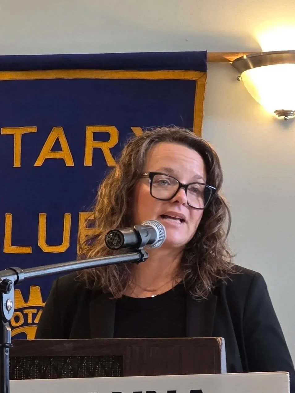 A woman with curly hair and wearing glasses speaking at a podium with a microphone, standing in front of a blue banner that says 'Rotary'.