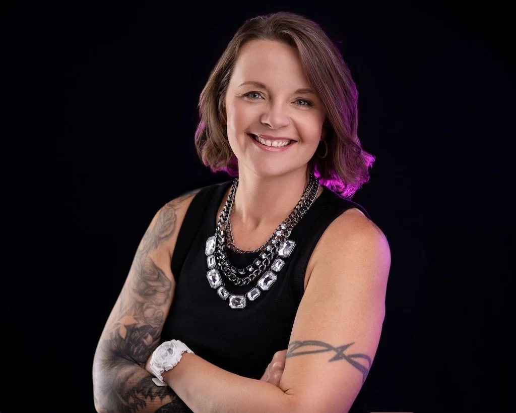 A smiling woman with short brown hair wearing a black sleeveless top, layered silver necklaces, a white wristwatch, and tattoos on her arms, posed against a black background.