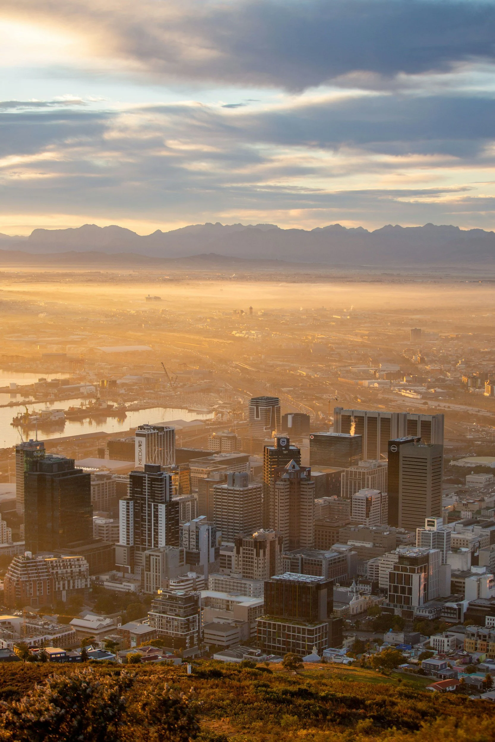 Aerial view of a city landscape at sunrise with tall buildings in the foreground and mountains in the background, with sunlight casting a golden glow.