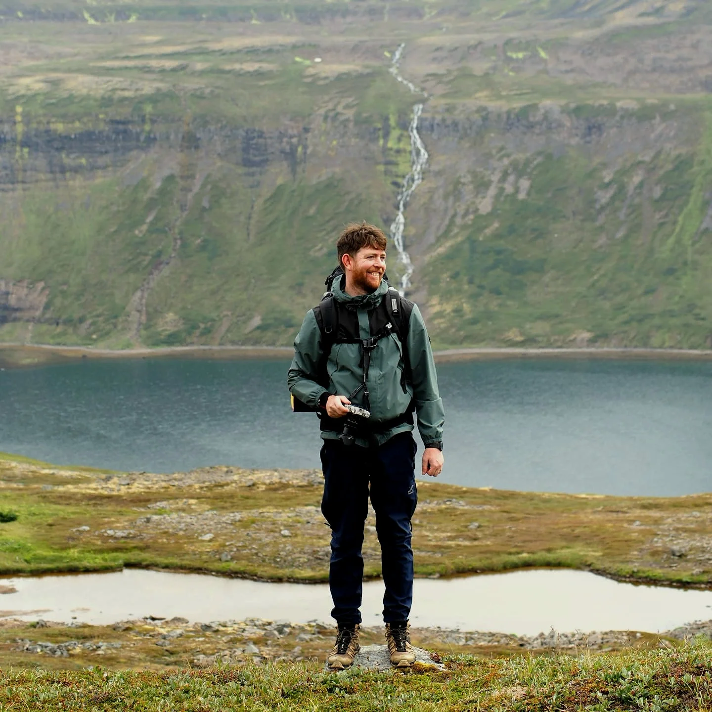 A man hiking outdoors near a lake with a mountain and waterfall in the background.
