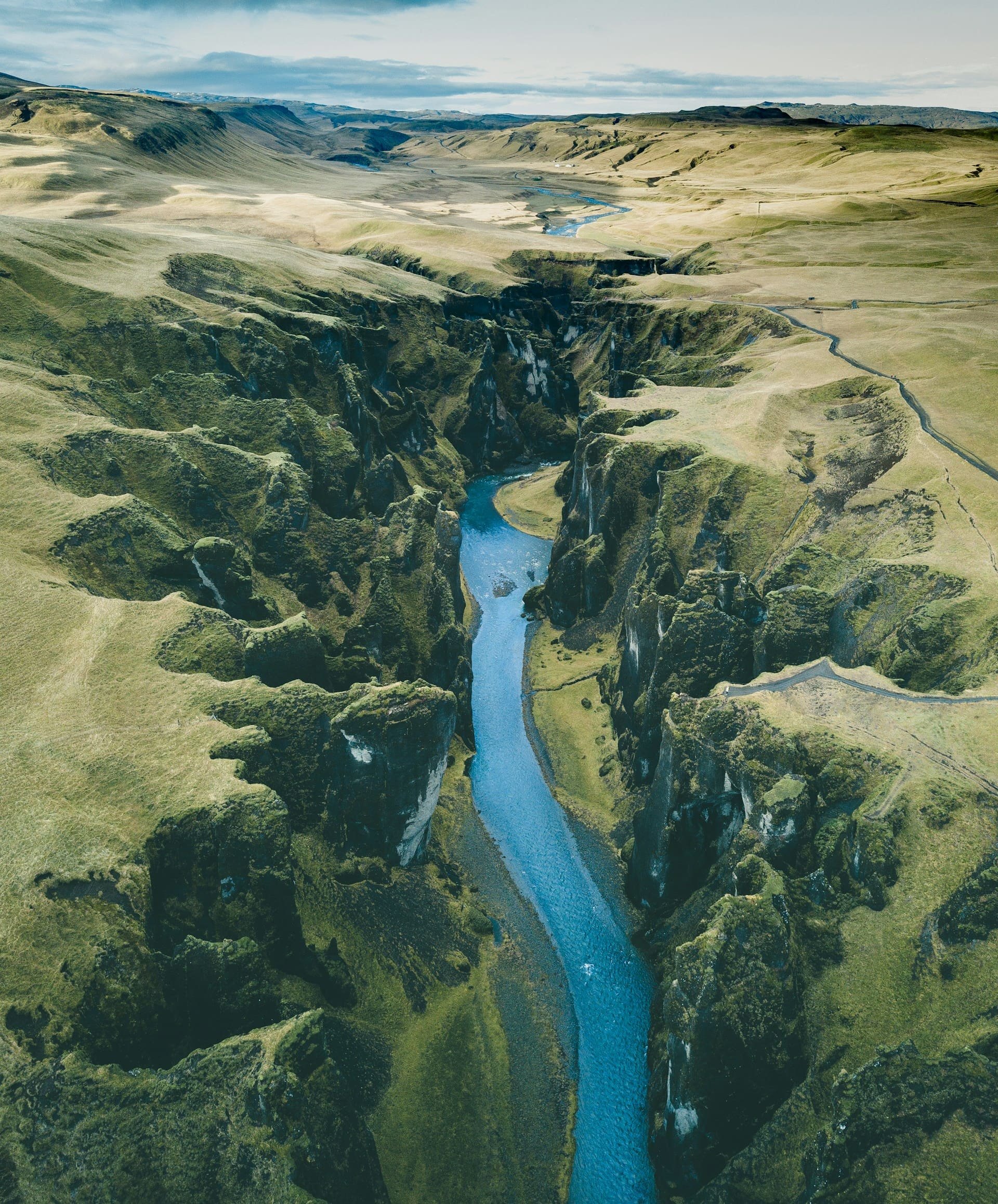 Aerial view of a canyon with a river running through it, surrounded by grassy hills and cliffs under a cloudy sky.