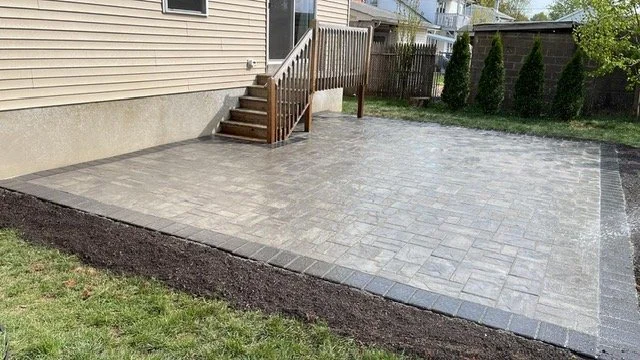 Newly paved backyard patio with concrete pavers, adjacent to a house with stairs leading to a door, surrounded by grass and a wooden fence with growing trees.