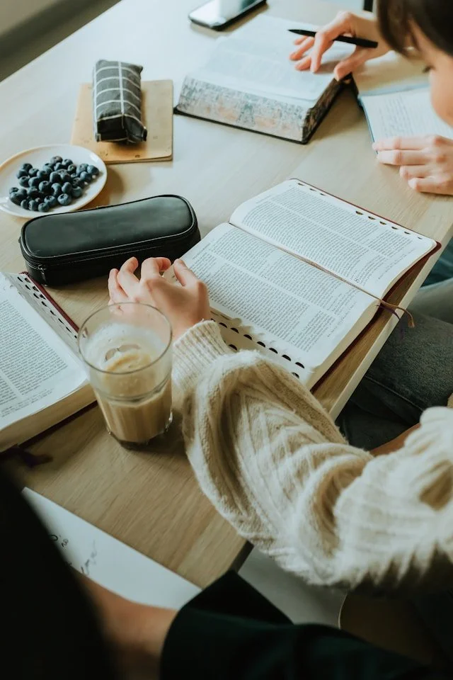 People studying together at a table with books, a bowl of blueberries, a glass of iced coffee, and stationery.