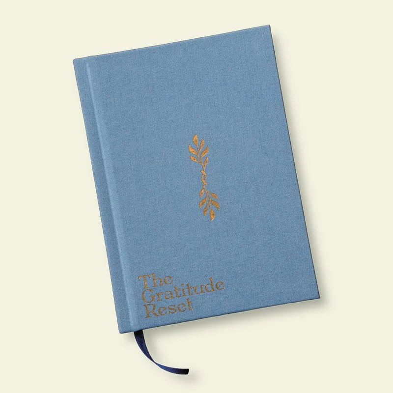 A light blue hardcover journal titled "The Gratitude Reset" with a gold foil leaf design on the cover and a black ribbon bookmark.