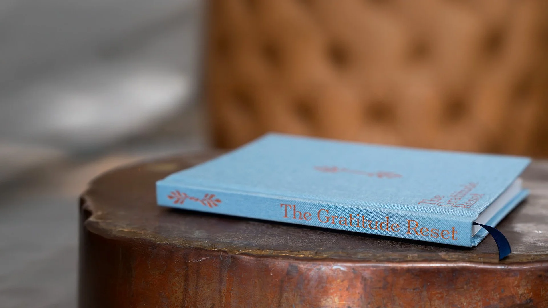 A blue book titled "The Gratitude Reset" rests on a round wooden table with a textured surface. In the background, a blurred leather or faux leather chair is visible.