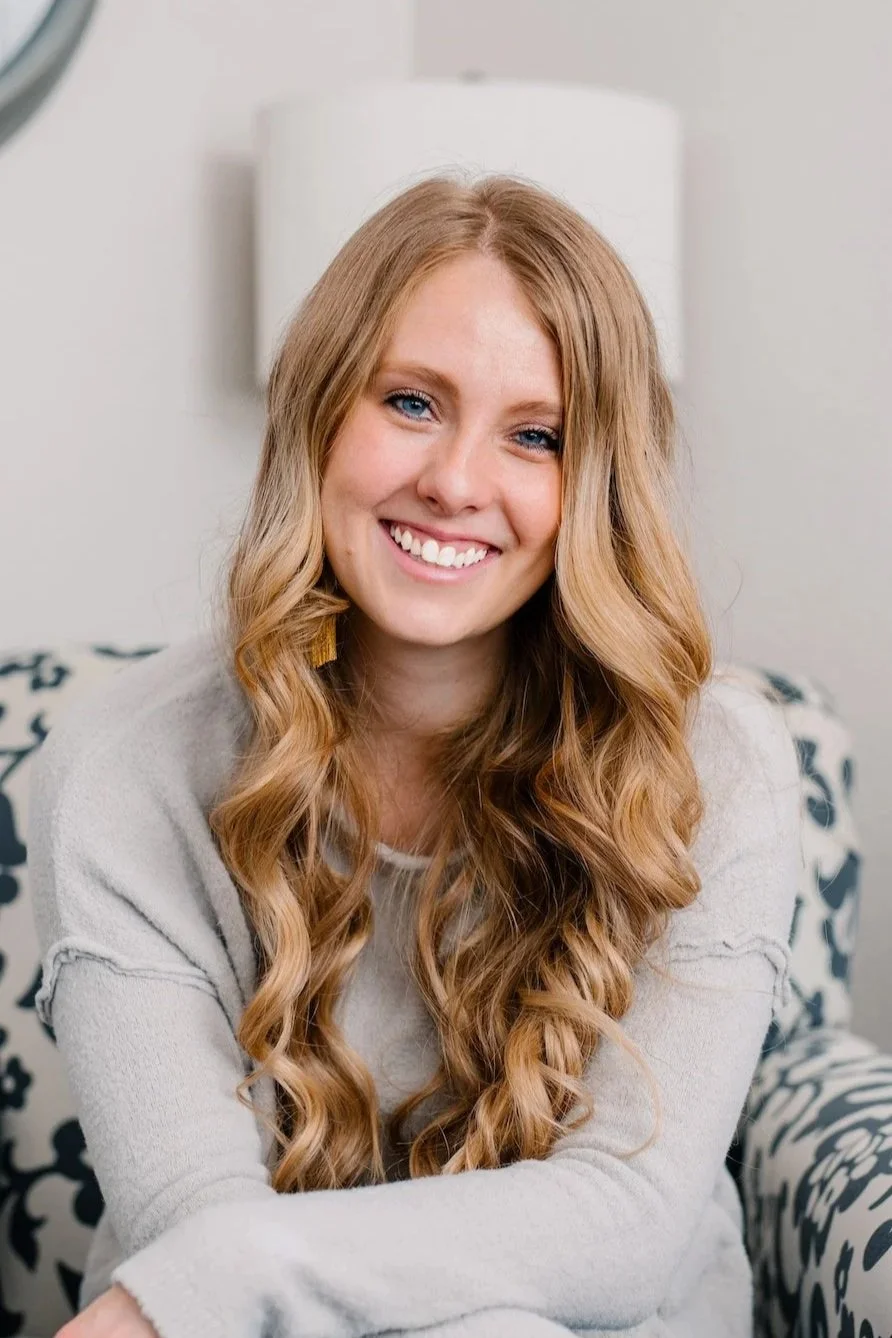 A young woman with long, wavy red hair, smiling and sitting on a black and white patterned sofa in a cozy, well-lit room.