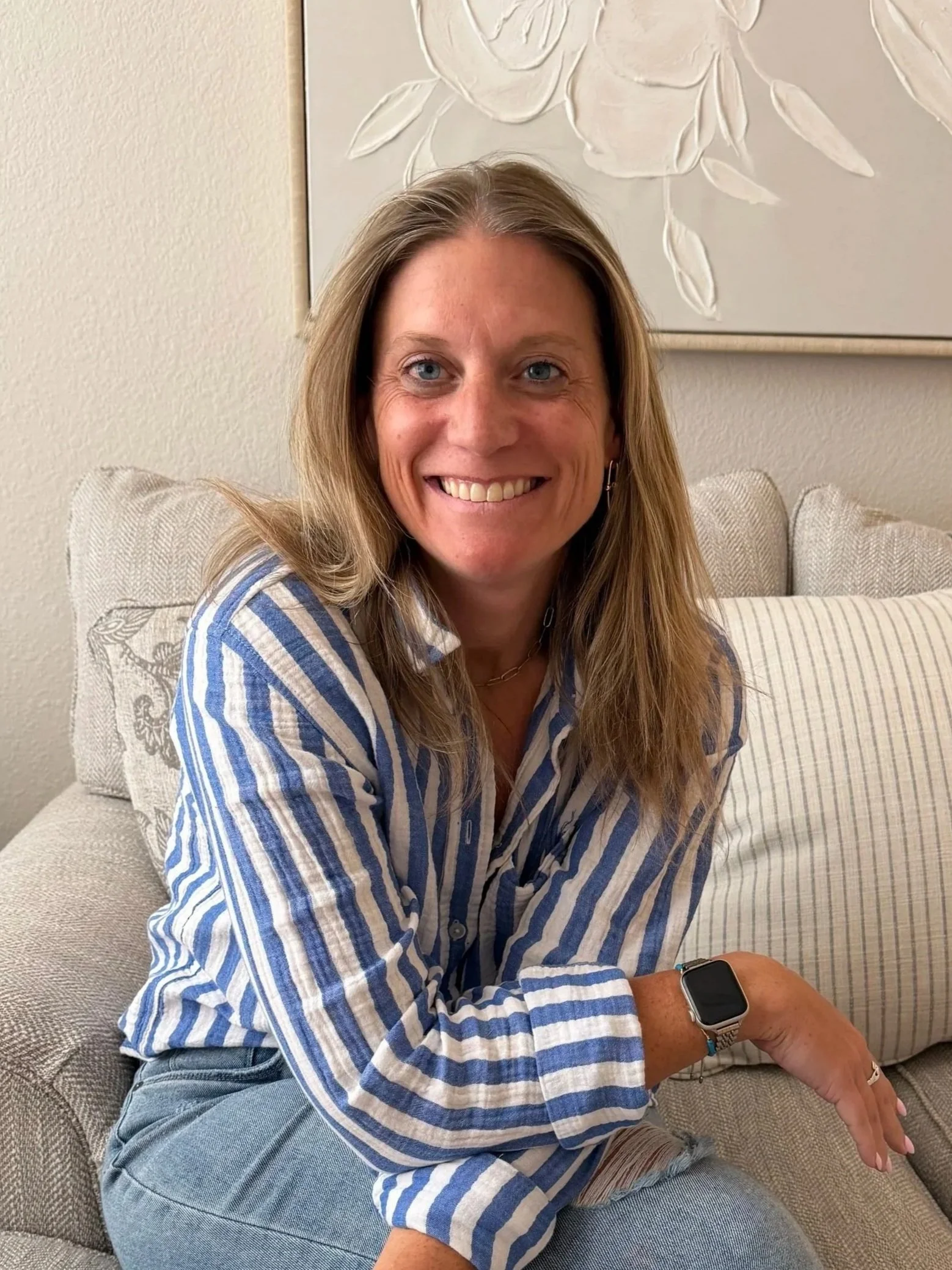 A woman with long light brown hair and blue eyes sitting on a beige couch, smiling. She is wearing a blue and white striped shirt and has a smartwatch on her wrist. There is a white piece of wall art with a floral design behind her.