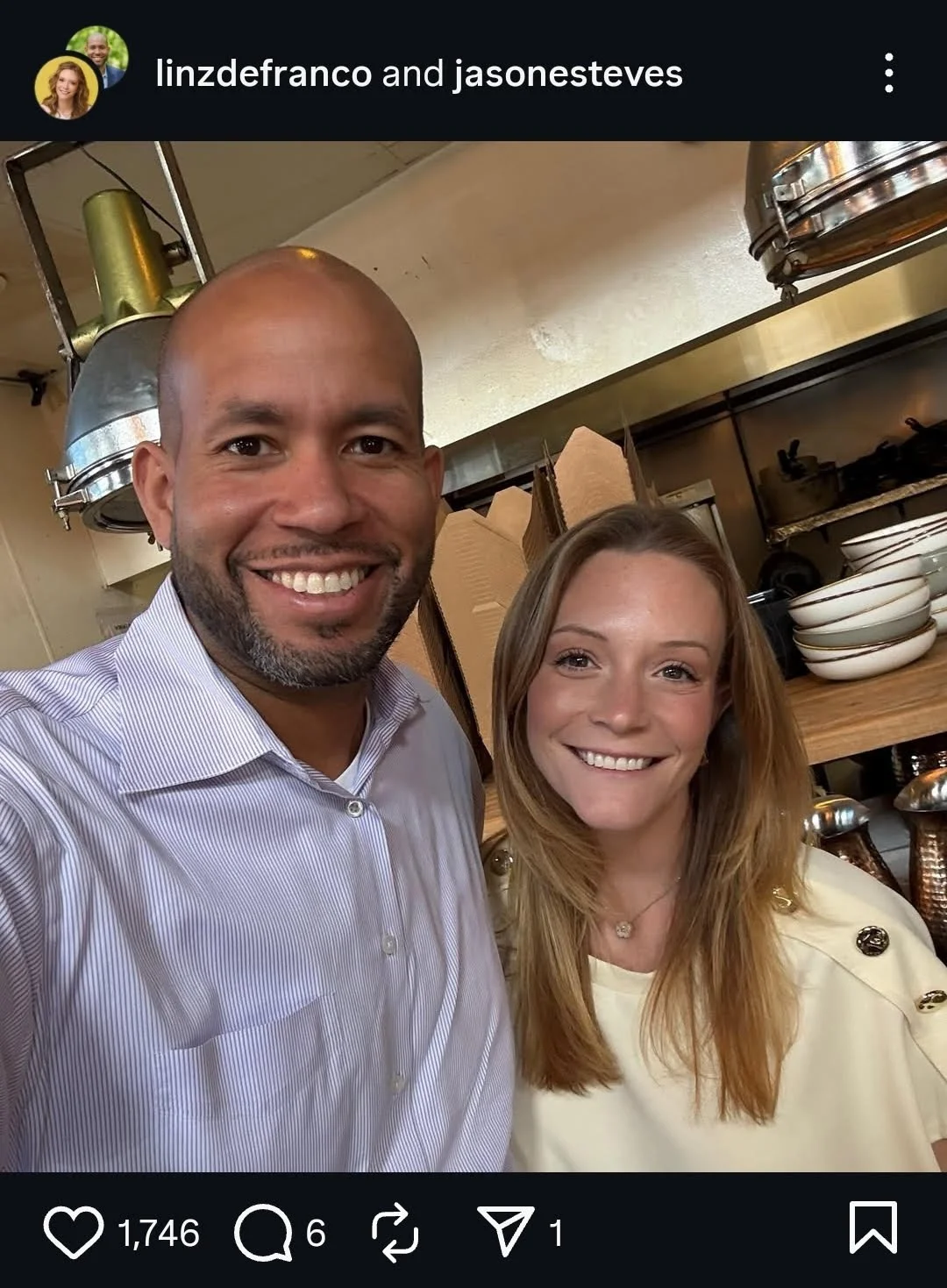 A man and a woman smiling for a selfie in a kitchen or restaurant setting, with dishes and kitchen equipment in the background.