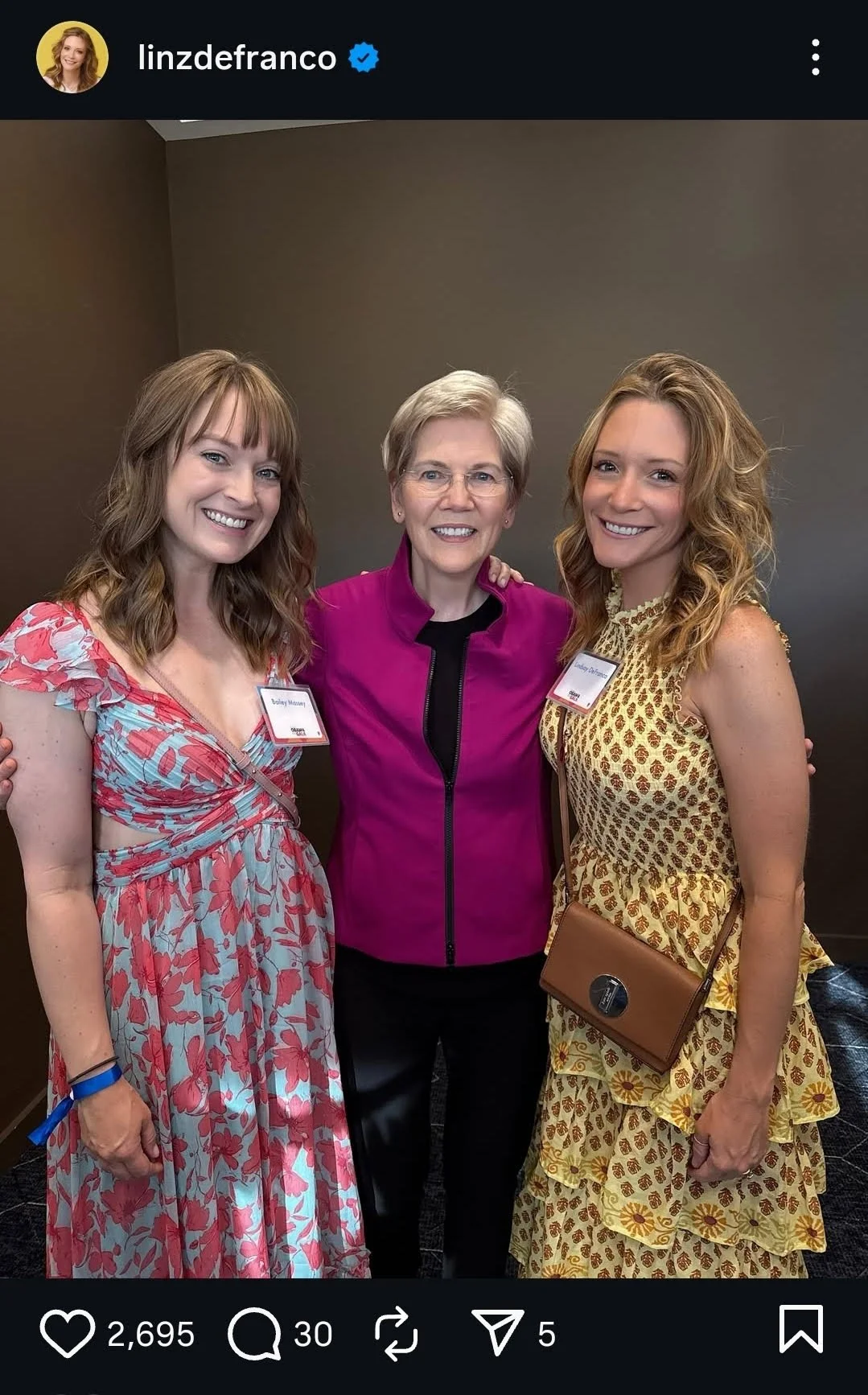 Three women standing together at an event, smiling at the camera. Sen. Elizabeth Warren is in the middle wearing a magenta blazer and glasses, with her arms around two women in floral dresses, they are Lindsay DeFranco and a friend