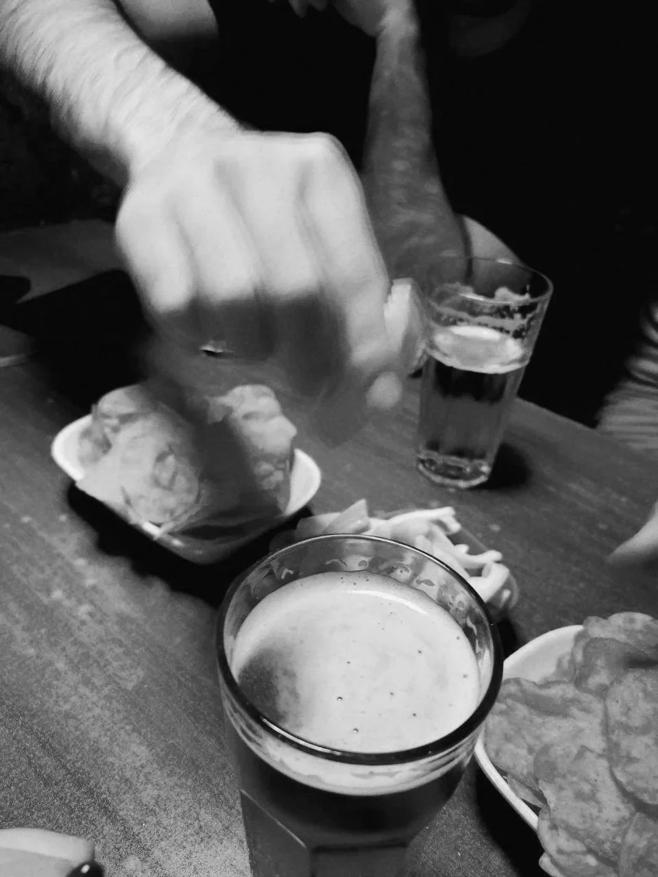Black and white photo of a table with a glass of beer, a bowl of fried food, and a person holding a spoon with food.