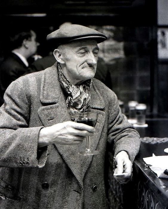 An elderly man holding a glass of red wine in a vintage-style setting, dressed in a tweed coat and a beret, with a confident expression.