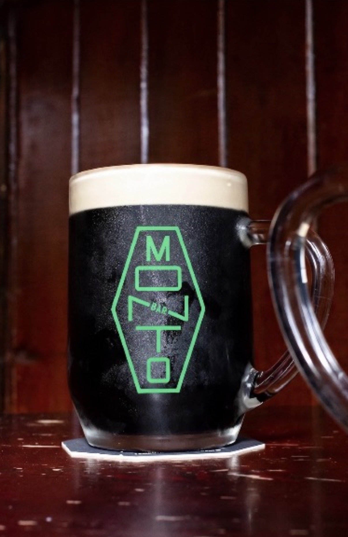 A glass beer mug with foam on top, featuring a green logo with the words 'MOTO BAR' on a dark background, placed on a wooden surface.