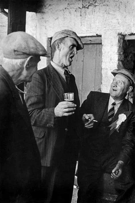 Three men wearing caps and suits, engaging in conversation, one holding a glass, indoors with wooden wall background.
