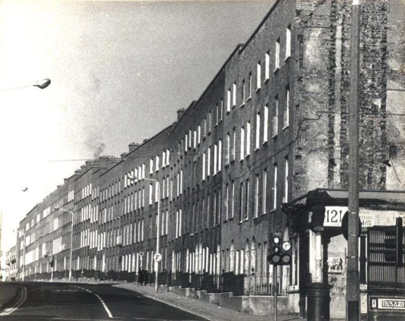 A black-and-white photo of a street with multi-story apartment buildings, a streetcar stop with a shelter, streetlights, and utility poles along the roadway.