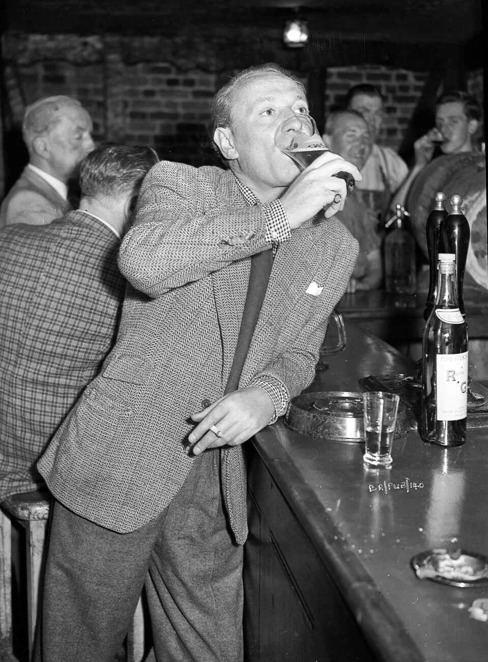 A man in a checkered blazer and tie is leaning against a bar while drinking beer from a pint glass in a lively pub with other men socializing in the background.