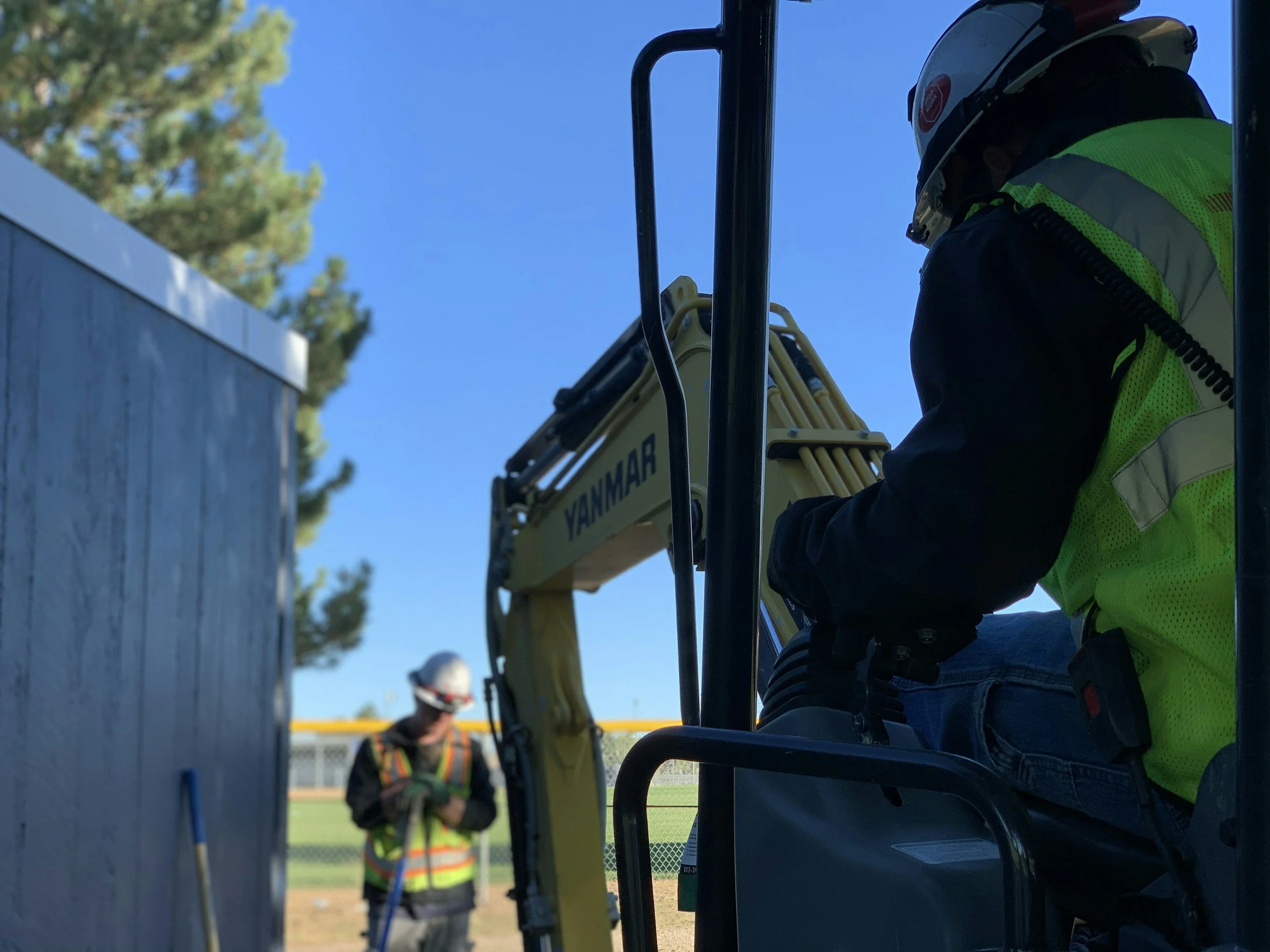 Construction worker operating a small backhoe excavator on a sunny day, with another worker in the background near a fence.