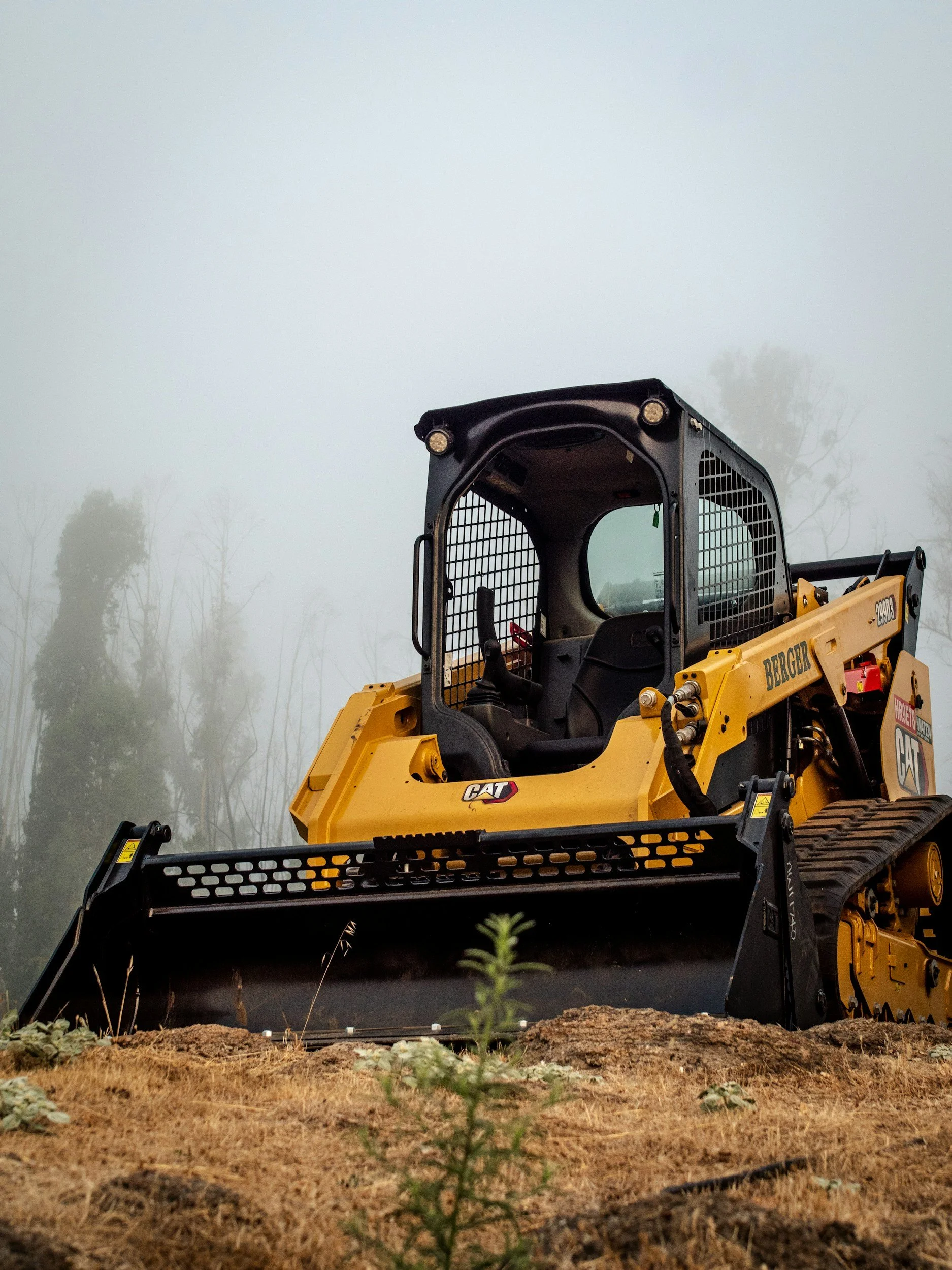 A yellow construction bulldozer with black accents working on a foggy outdoor site, surrounded by dirt and some plants.