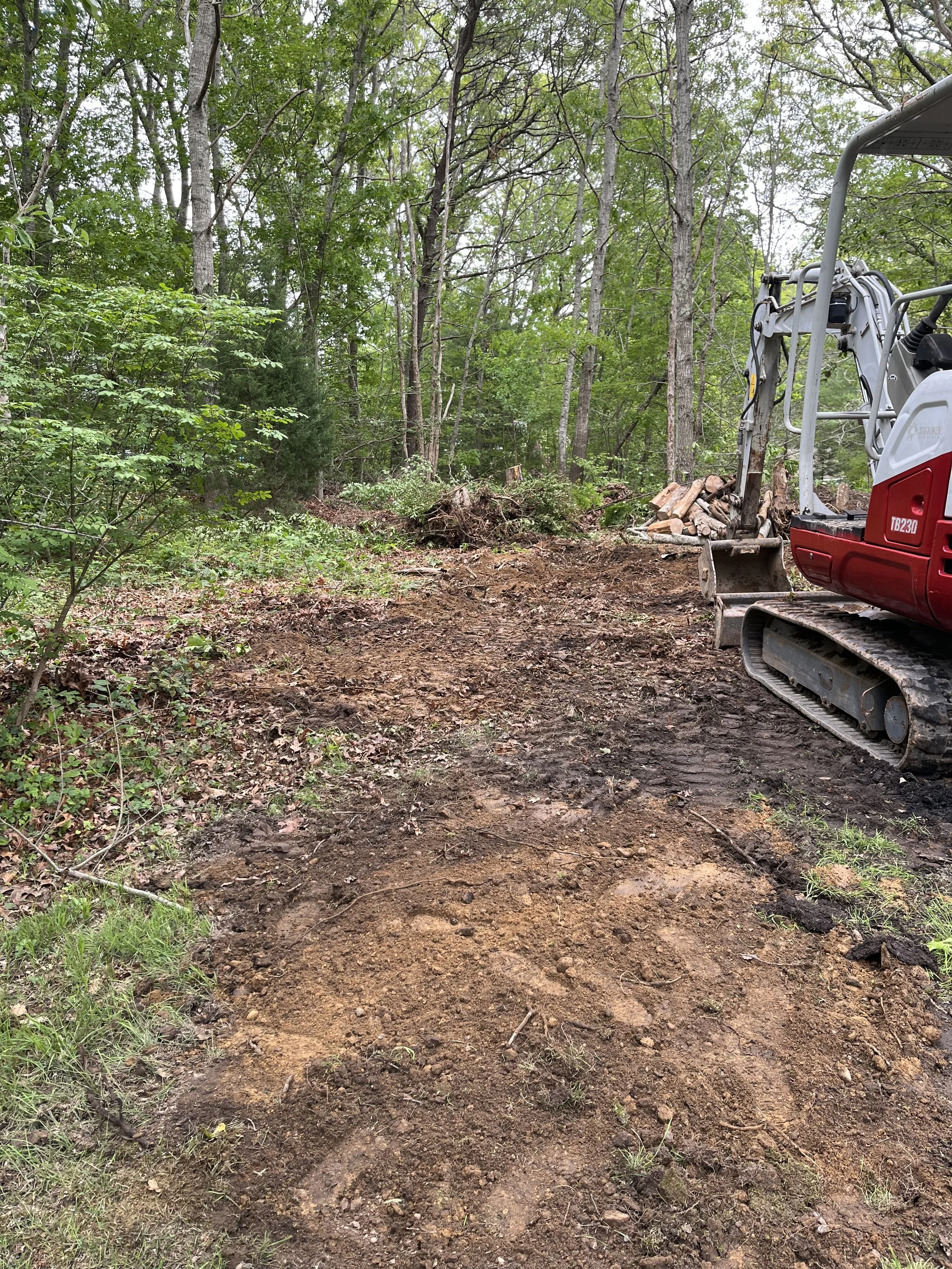 A dirt clearing in a wooded area is being prepared with a small excavator. There are trees with green foliage in the background, and a pile of logs on the right side of the excavator.