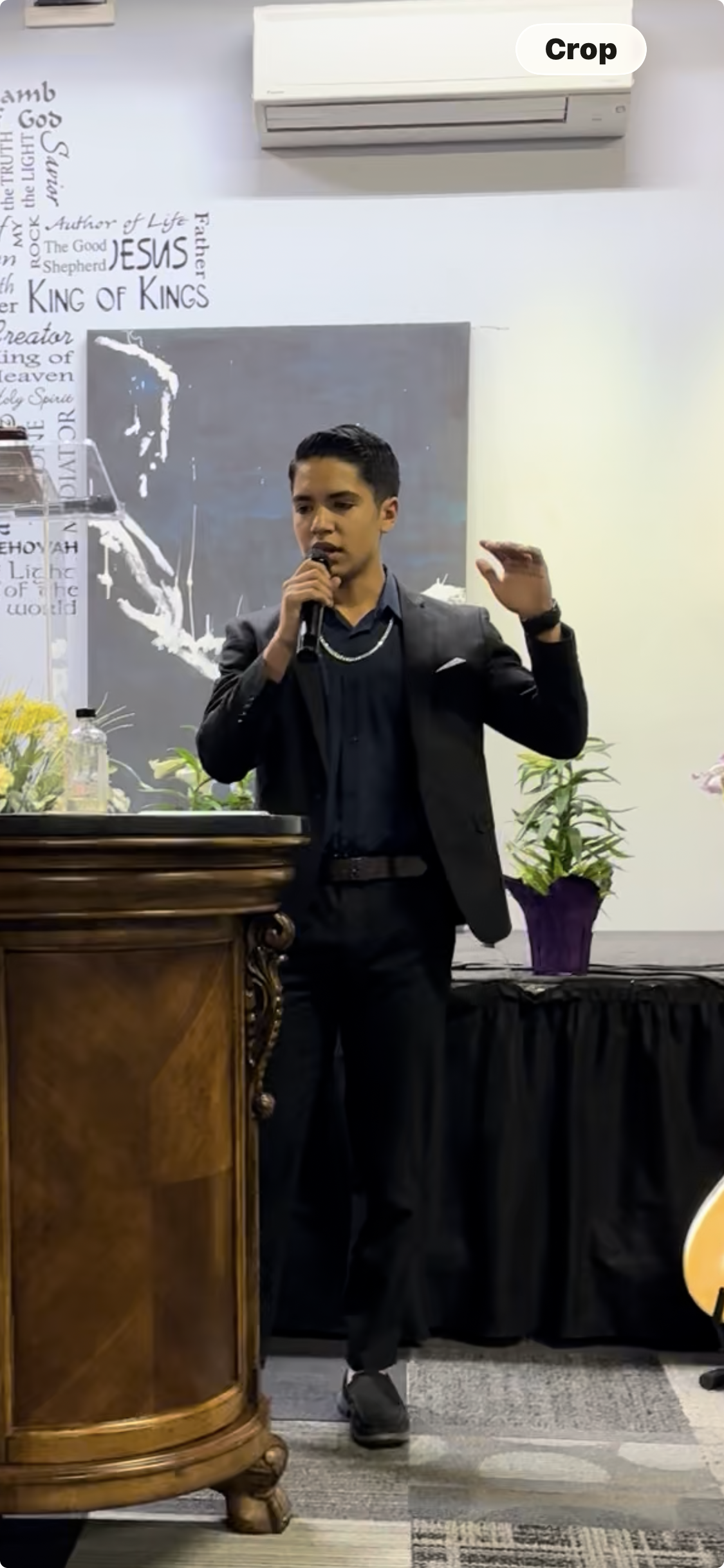 Nehemiah Castillo in a business suit speaking into a microphone at an event, standing behind a wooden podium with a bottle of water and some flowers. There is a blackboard and potted plants in the background, and an air conditioning unit on the wall.