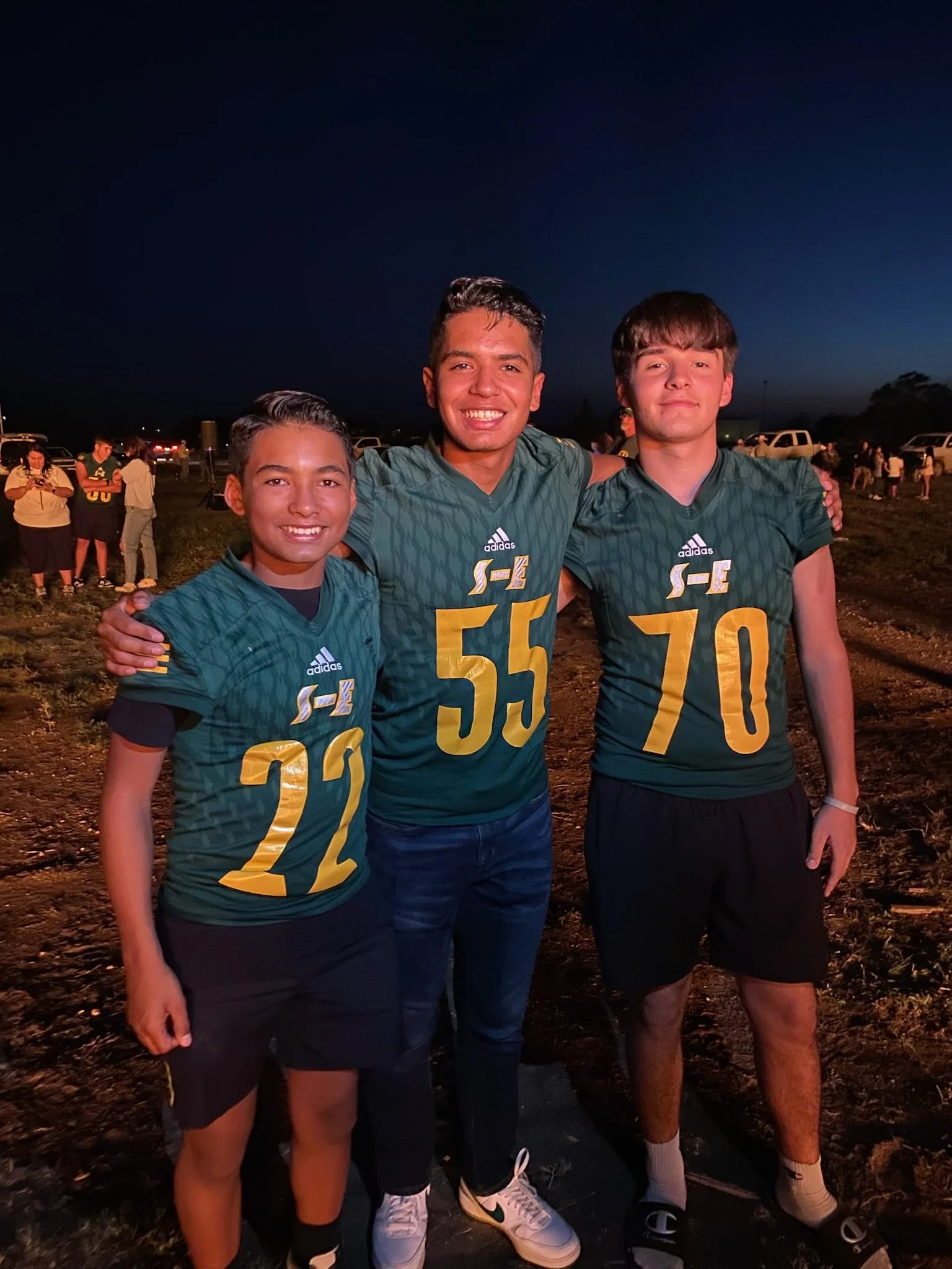 Nehemiah Castillo and two others in green jerseys with yellow numbers and the Adidas logo, standing outdoors at night, smiling and posing with arms around each other's shoulders. The background shows a dark sky and other people and vehicles.