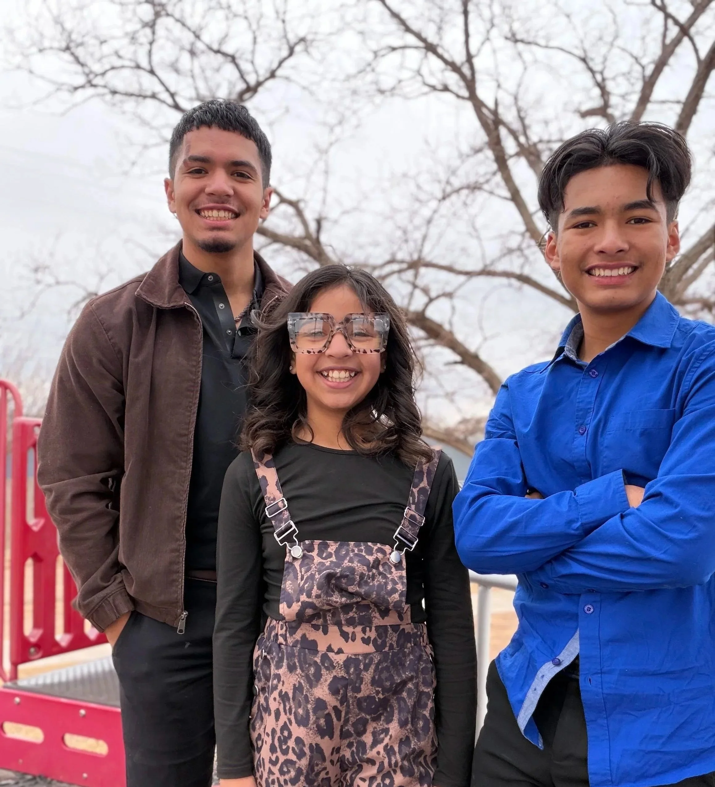 Nehemiah Castillo with his two younger siblings smiling outdoors in front of leafless trees, including his sister in glasses and leopard print overalls, and Nehemiah and his younger brother, one in a brown jacket and the other in a bright blue shirt.