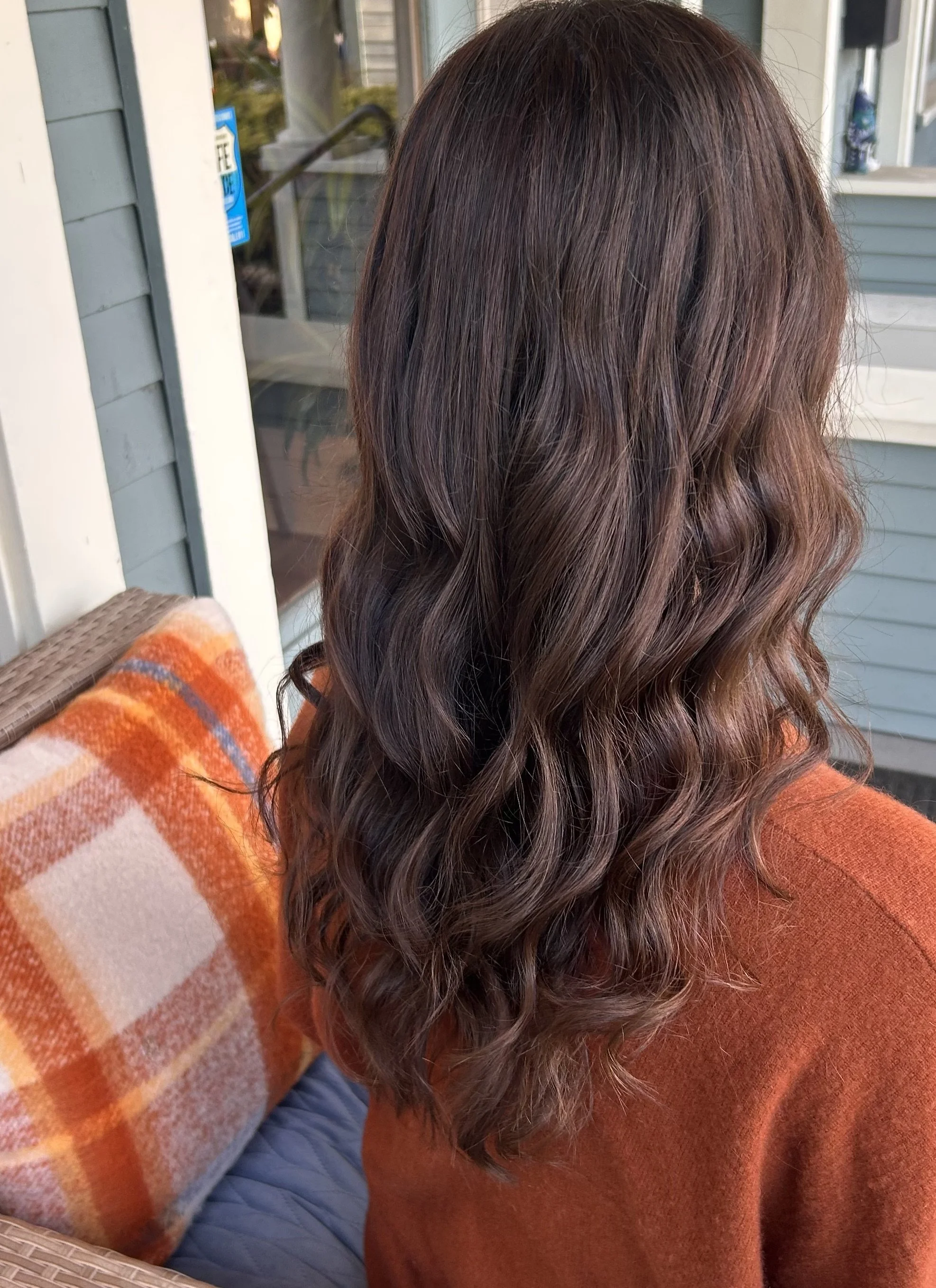 Back view of a person with long, wavy dark brown hair wearing an orange sweater on a porch